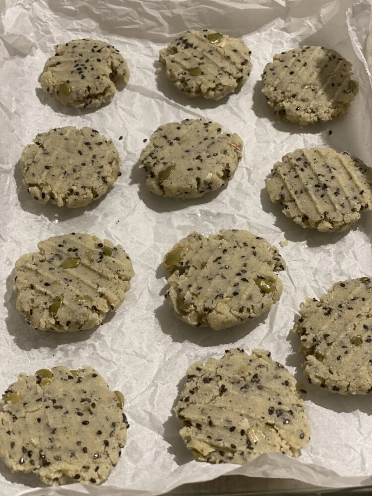 Flattened cookie dough rounds with black sesame seeds and pumpkin seeds on parchment paper, ready for baking.