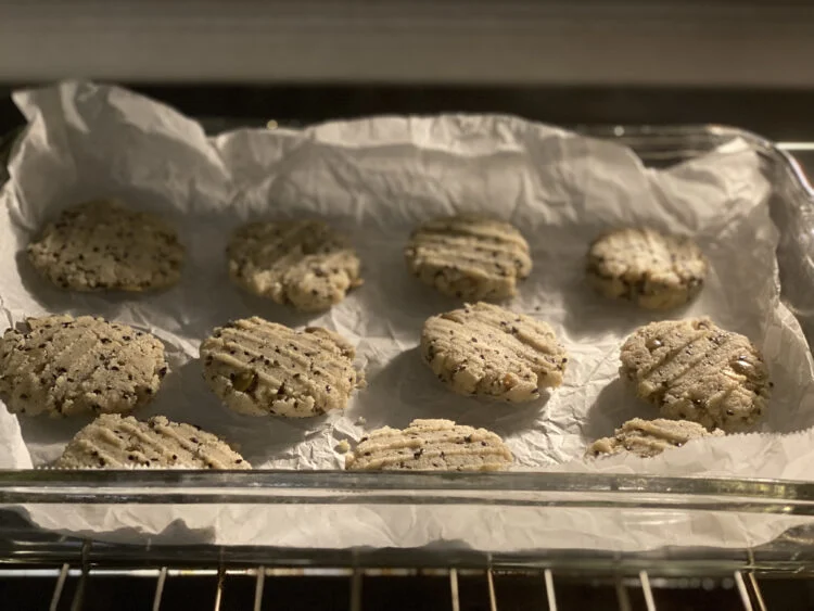 Flattened cookies with black sesame seeds and pumpkin seeds on parchment paper in a glass baking dish, placed in the oven.