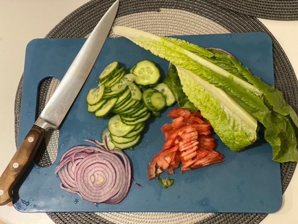 Fresh burger toppings on a blue cutting board: sliced cucumbers, red onions, diced tomatoes, and crisp lettuce.