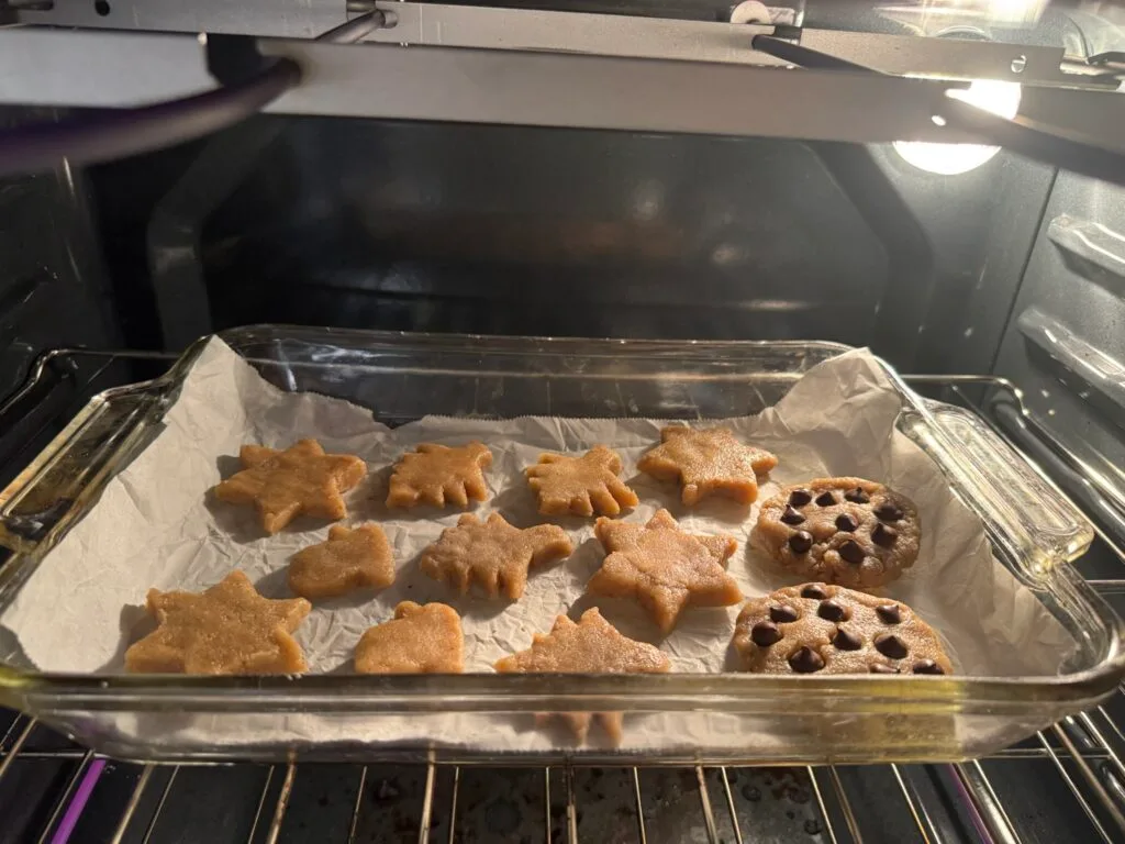Gluten-free Christmas cookies shaped like stars and other festive designs baking in the oven on a parchment-lined tray.