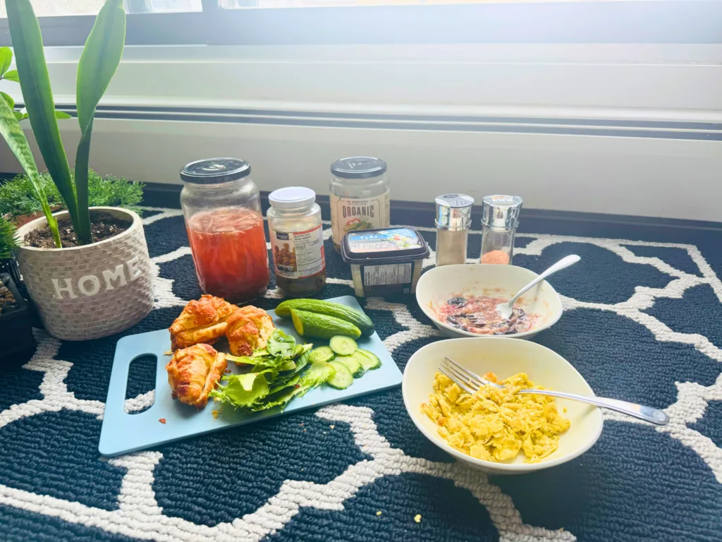 Mini croissants, fresh greens, cucumbers, and scrambled eggs on a cutting board with condiments and seasonings in the background.