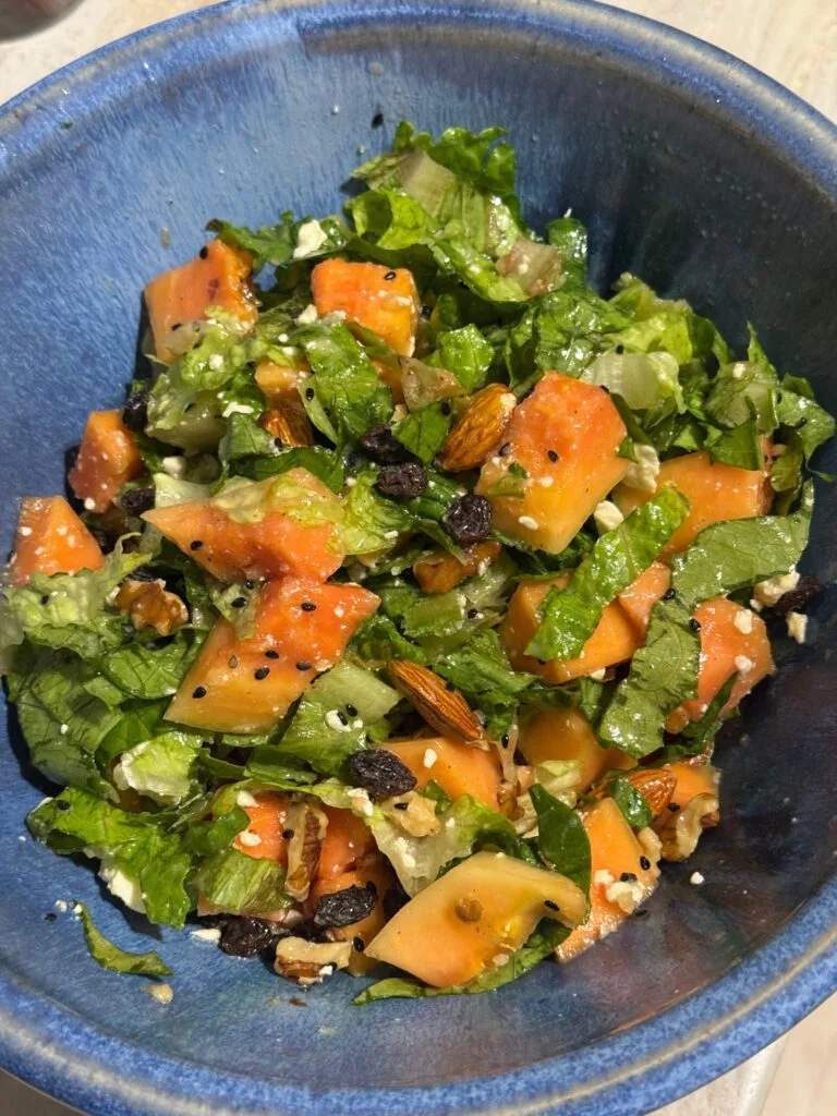 Bowl of papaya salad with lettuce, nuts, feta, and raisins.