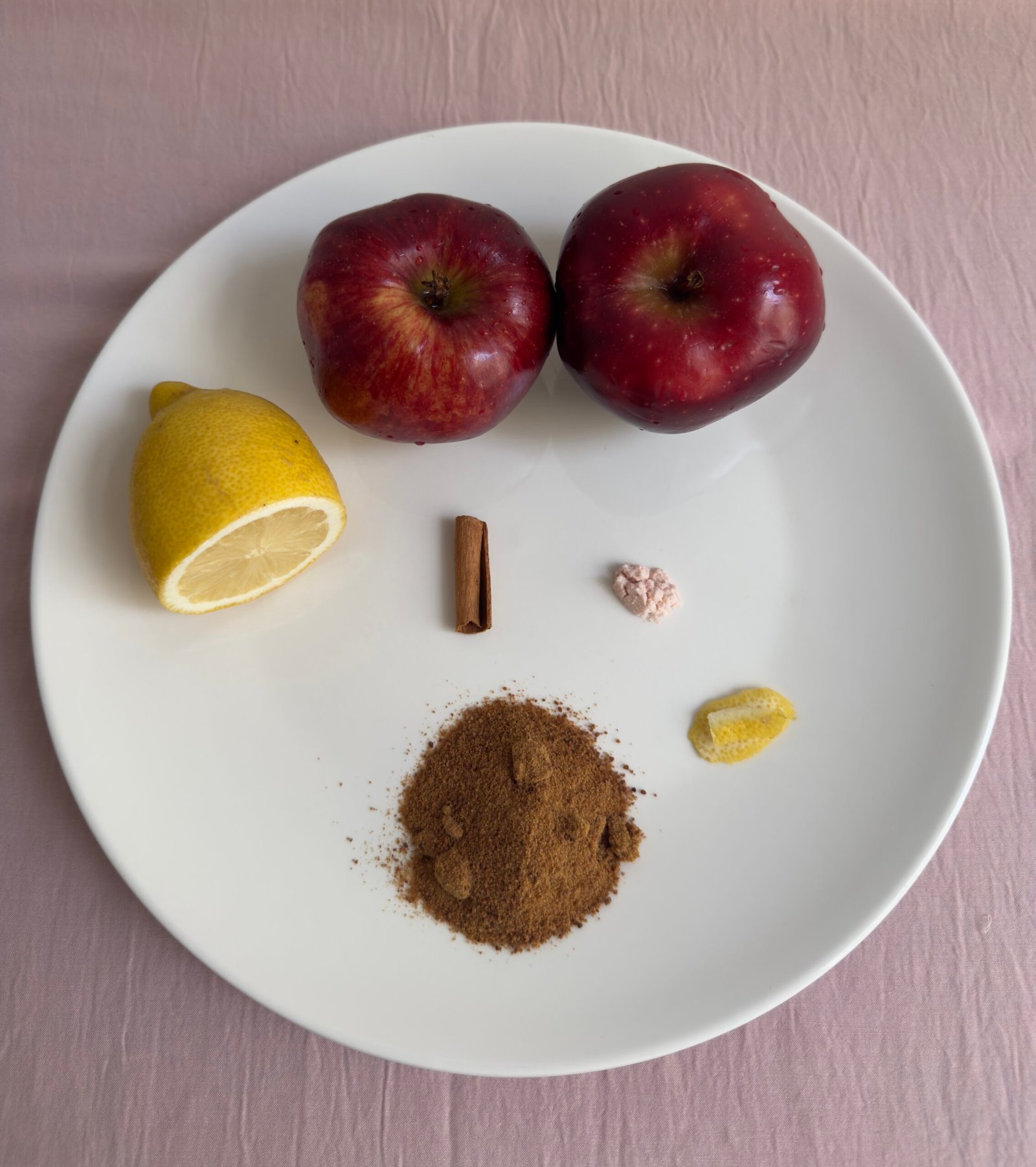 Flat lay of homemade applesauce ingredients on a white plate including two red apples, a halved lemon, a Ceylon cinnamon stick, coconut sugar, and a strip of lemon peel on a pink background