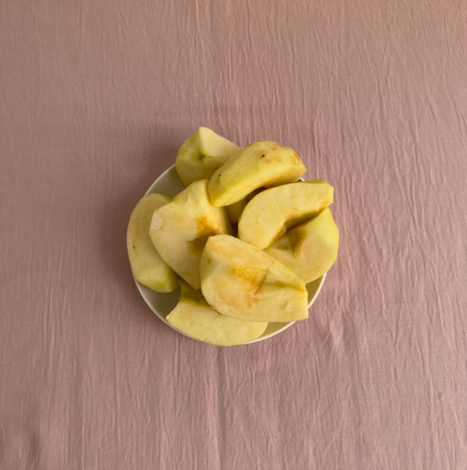 Peeled and quartered apple chunks in a small white bowl on a pink surface ready for cooking
