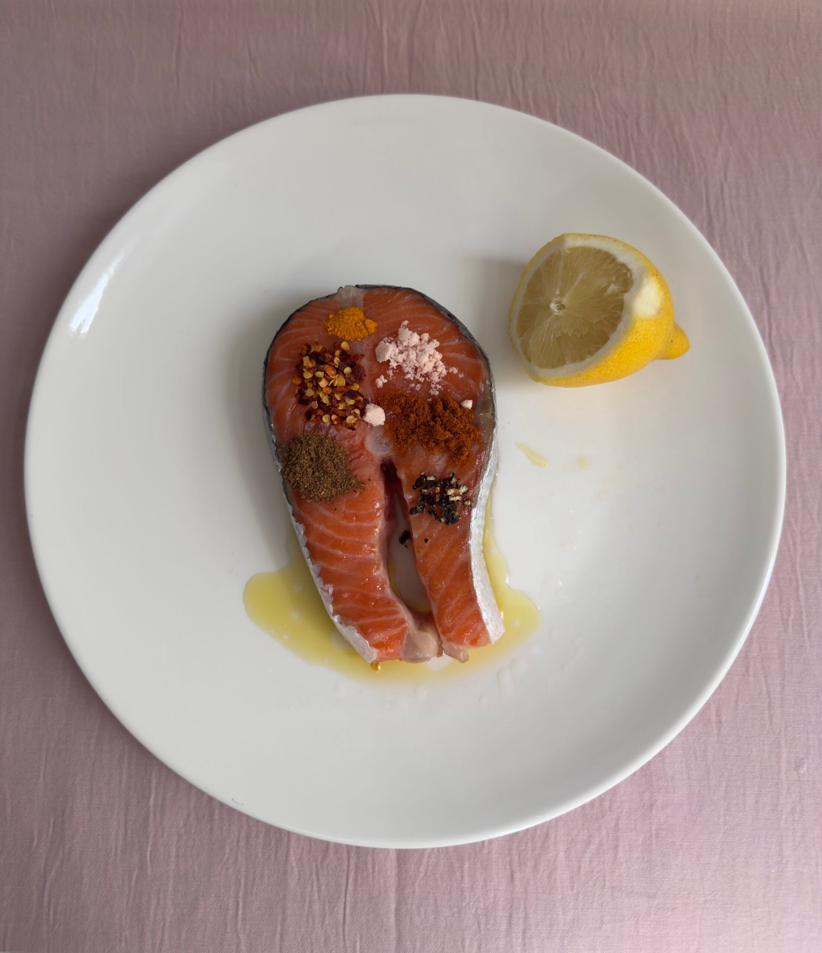 Raw salmon steak with dry spice rub (turmeric, paprika, crushed red chili, and salt) displayed on a white plate with a lemon wedge.