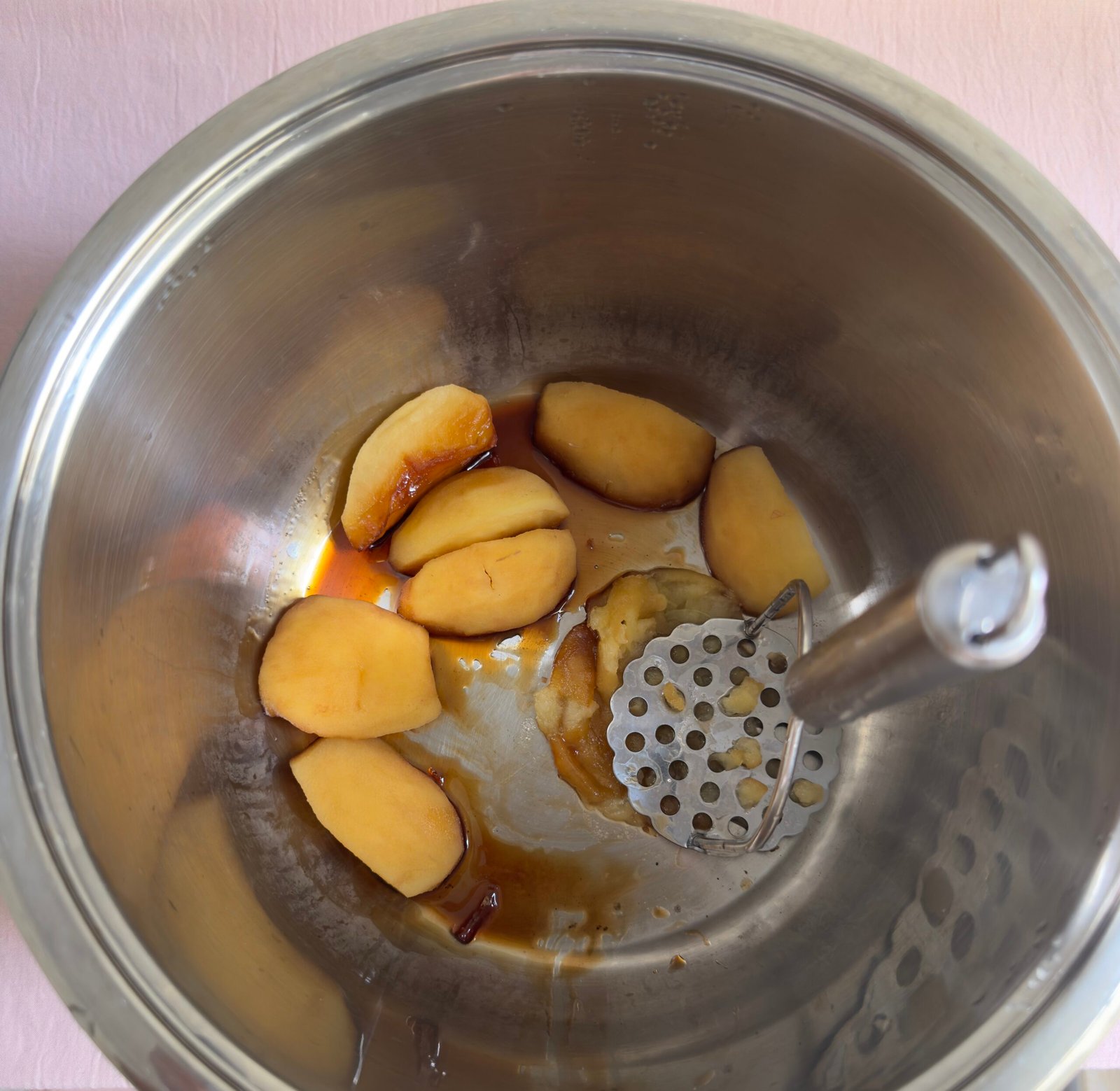 Fully cooked soft apple quarters in a stainless steel pot with a potato masher resting inside, ready to be mashed into applesauce