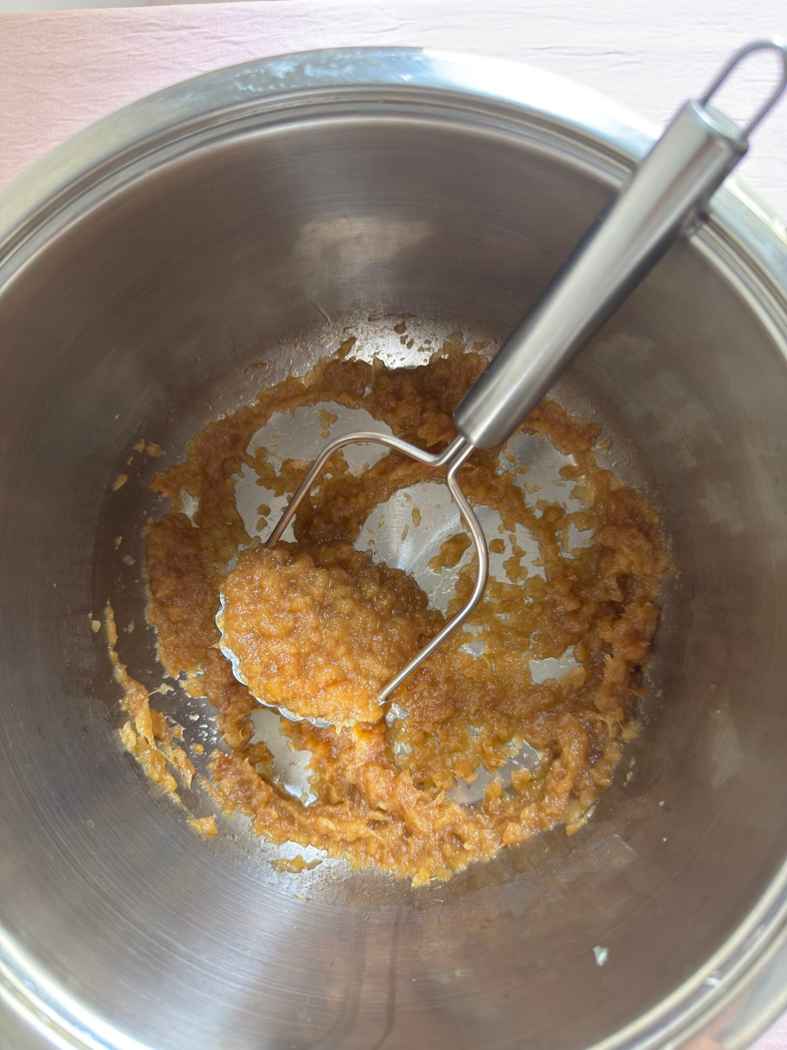 Freshly mashed homemade applesauce in a stainless steel pot with a potato masher, showing chunky golden texture mid-mash
