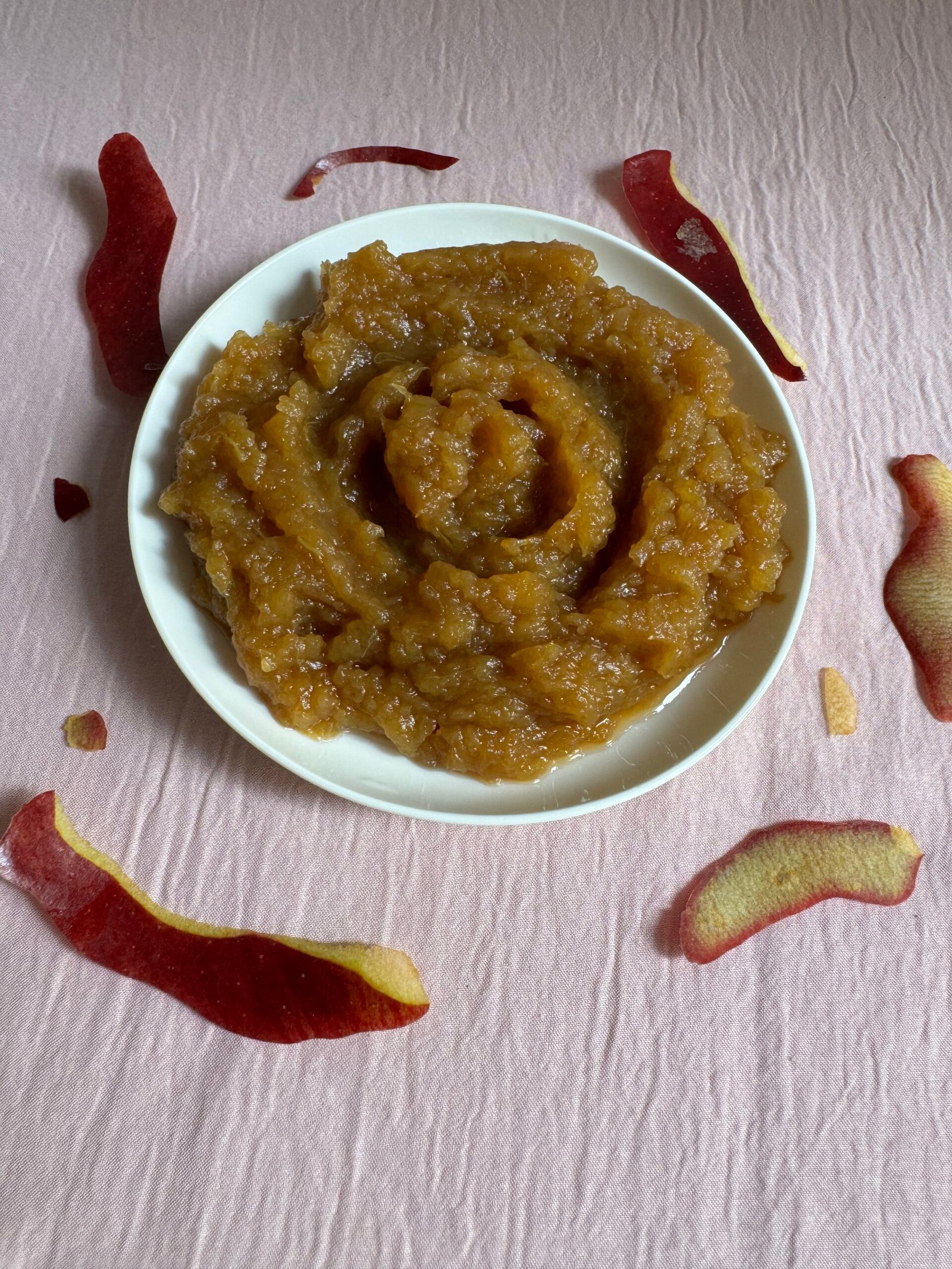 Homemade organic applesauce swirled in a white bowl surrounded by scattered red apple peels on a pink background