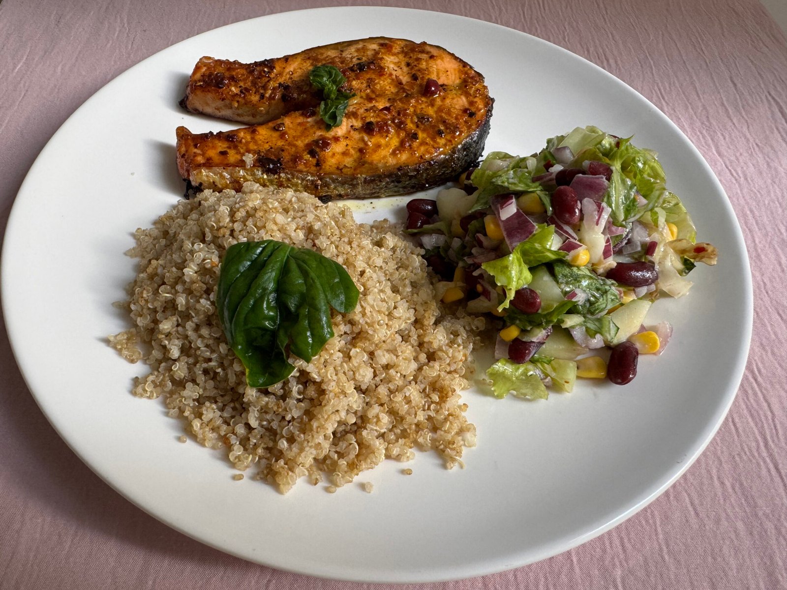 Finished plated meal with golden-brown baked salmon fillet topped with fresh basil, fluffy quinoa with basil leaf, and a side salad with red beans, cucumber, and corn.