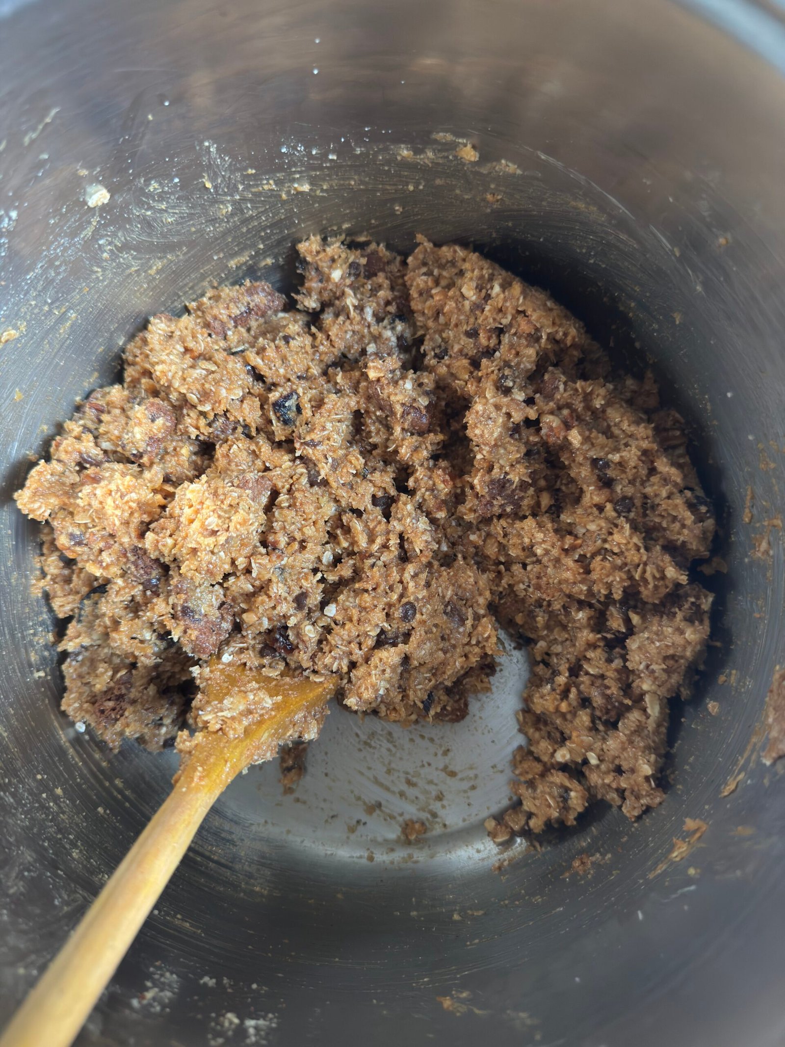 Gluten-free monster cookie dough mixed in a metal bowl with a wooden spoon, showing chunks of chocolate, raisins, and oats.