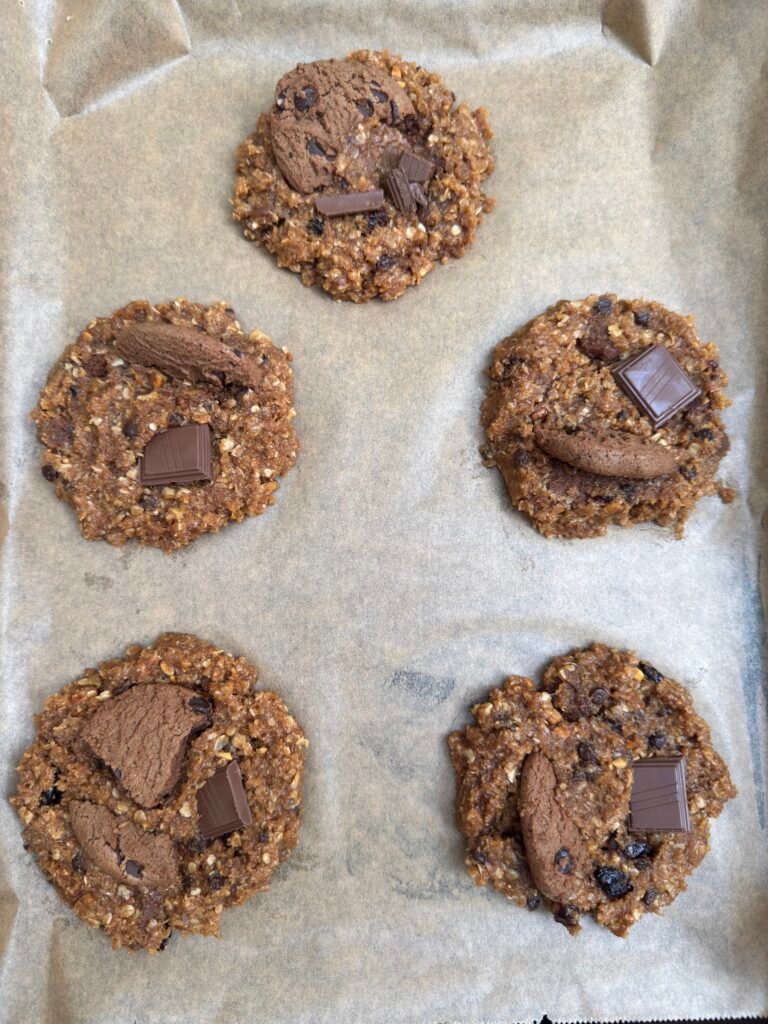 Five flattened monster cookie dough balls on a parchment-lined tray with chocolate chunks and cookie pieces pressed on top before baking.