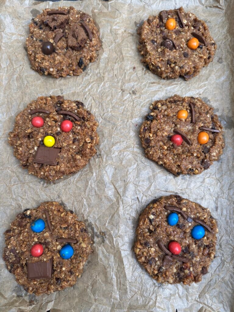Unbaked gluten-free monster cookies on parchment paper topped with colorful candies and chocolate pieces, ready for the oven.