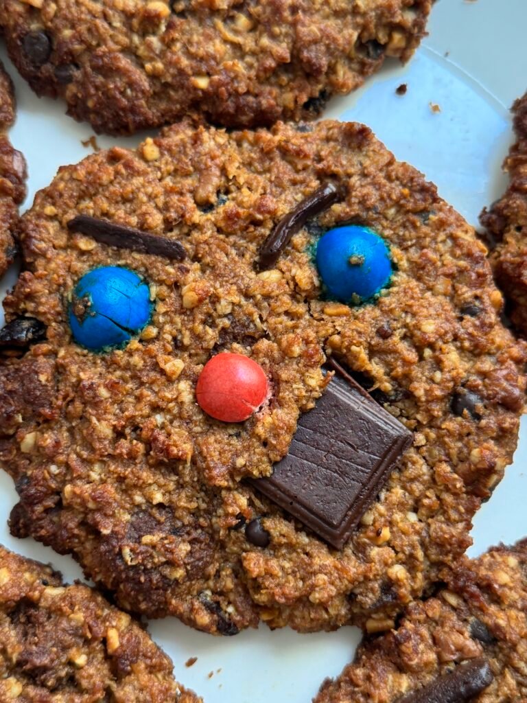 Close-up of a single gluten-free monster cookie with blue and red candies, dark chocolate squares, and visible oats and pecan chunks.