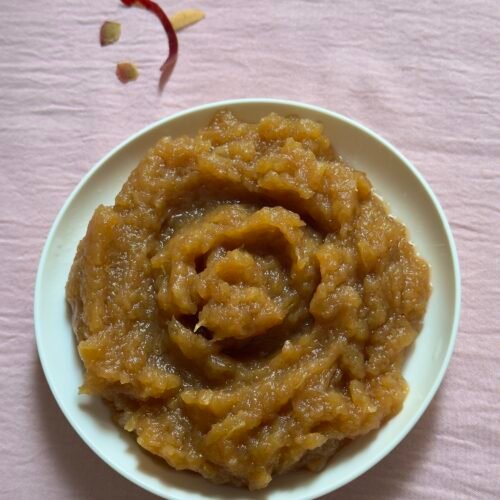 Homemade applesauce swirled in a small white bowl on a pink surface with a few apple peels scattered nearby