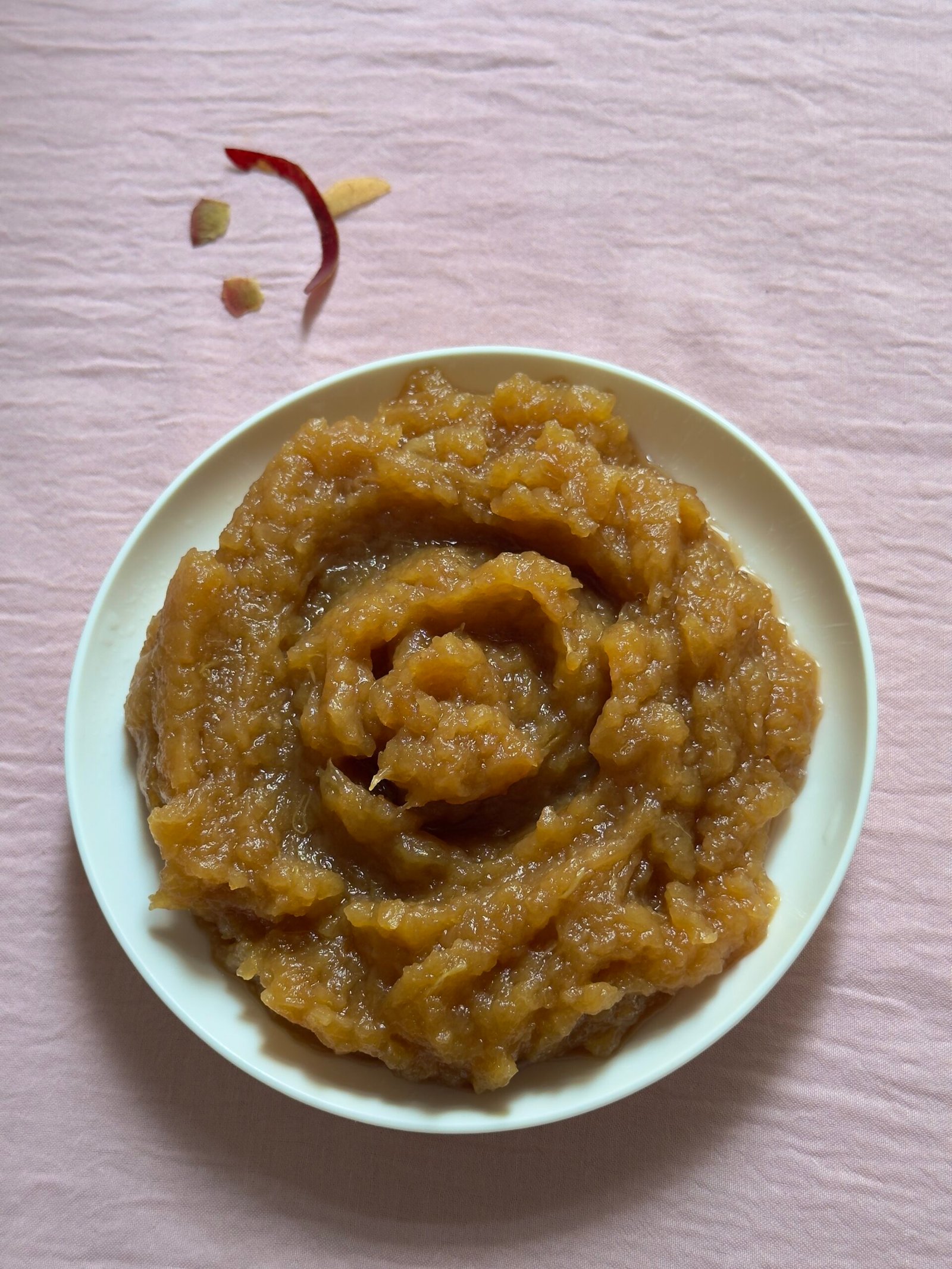 Homemade applesauce swirled in a small white bowl on a pink surface with a few apple peels scattered nearby