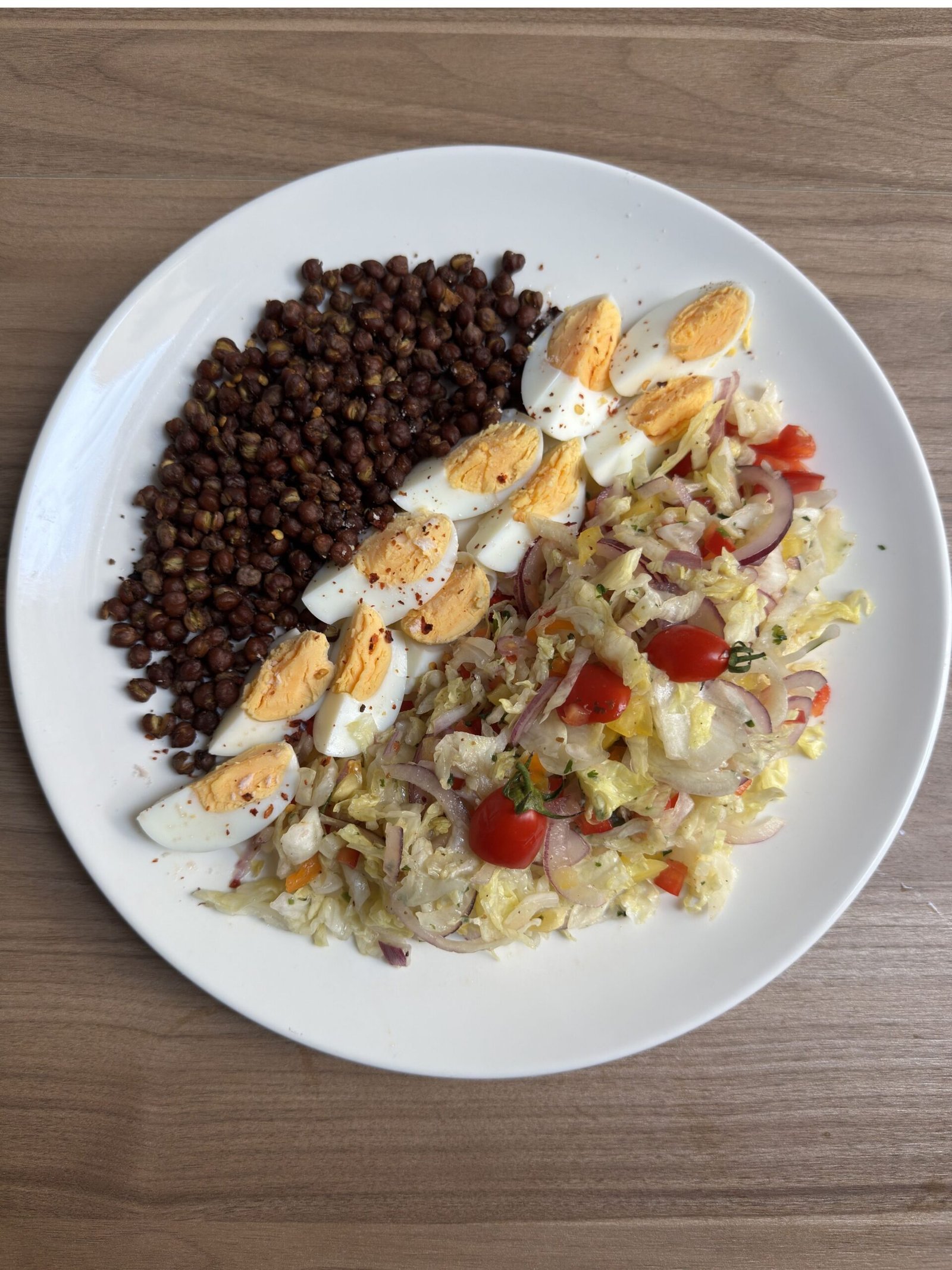 High-Protein Roasted Black Chickpea and Egg Salad plated on a white plate: roasted black chickpeas piled on the left, sliced hard-boiled eggs arranged in a line in the center, and dressed salad vegetables including chopped iceberg lettuce, red onion, cherry tomatoes, and mixed bell peppers on the right.
