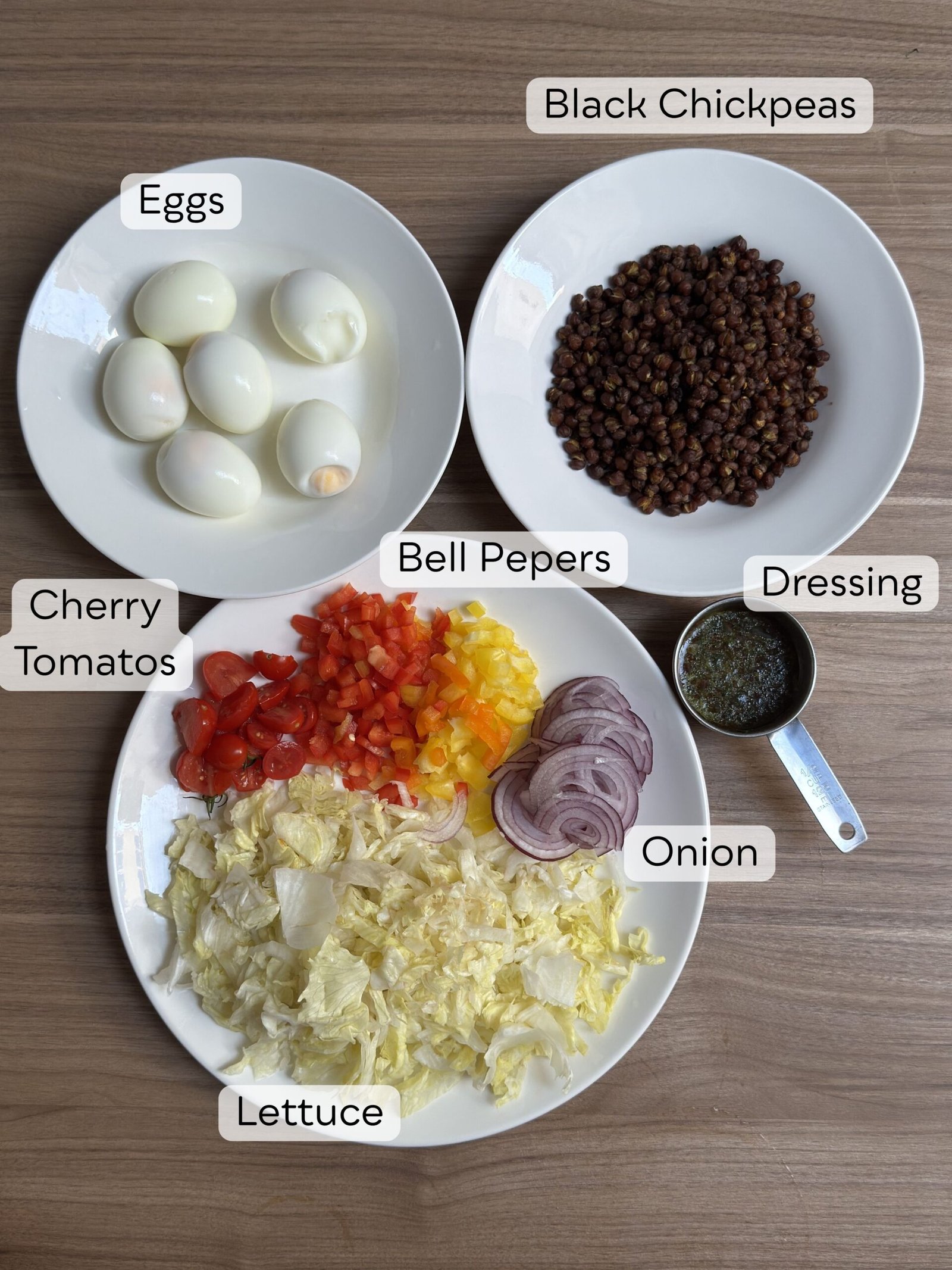 Overhead flat-lay of ingredients for High-Protein Roasted Black Chickpea and Egg Salad, displayed in separate labeled white bowls and plates: halved hard-boiled eggs, roasted black chickpeas, chopped red, orange, and yellow bell peppers, halved cherry tomatoes, sliced red onion, chopped iceberg lettuce, and honey-mustard dressing in a small measuring cup.