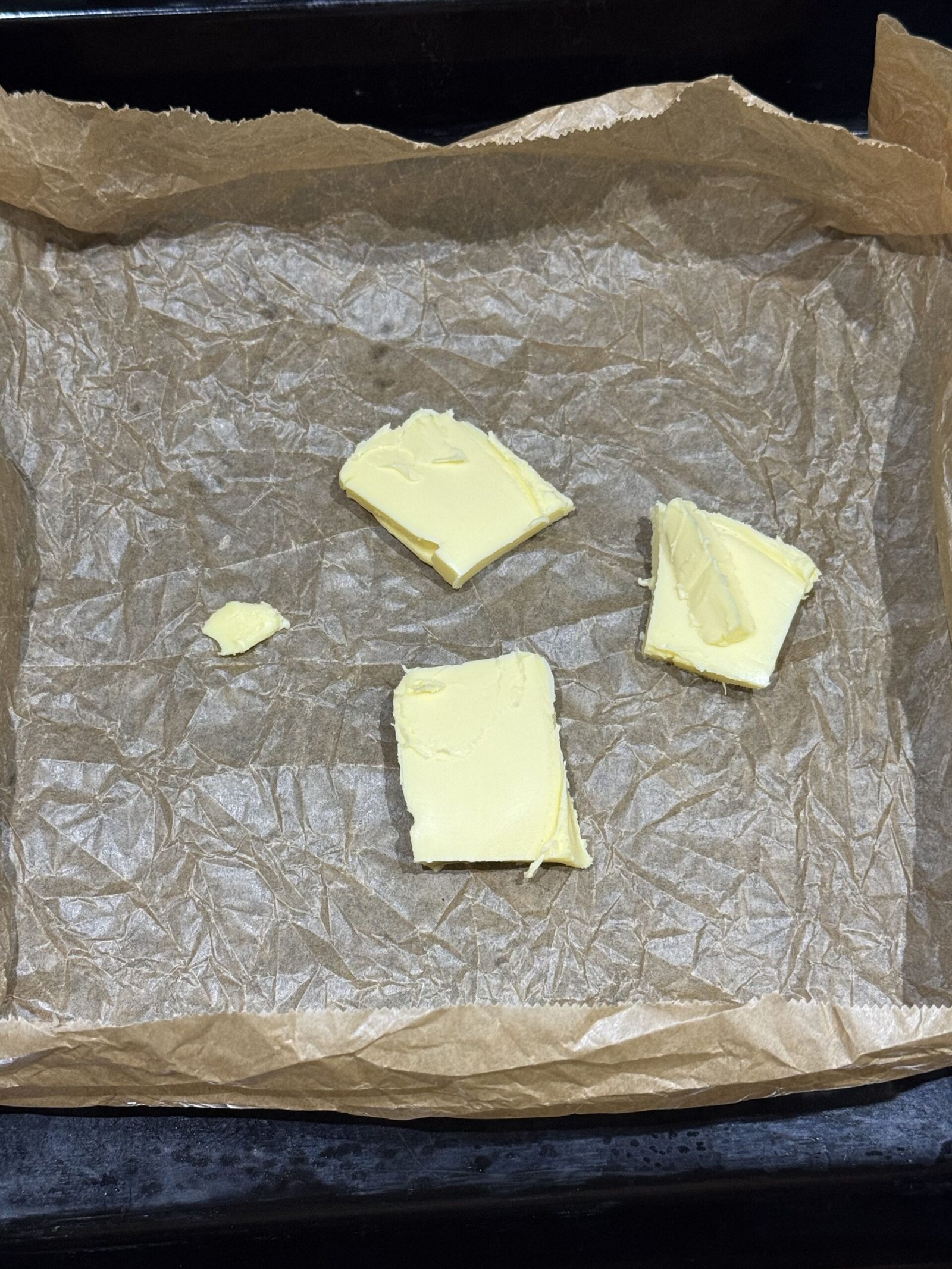 Sliced butter pieces placed on baking paper inside a black baking tray before melting