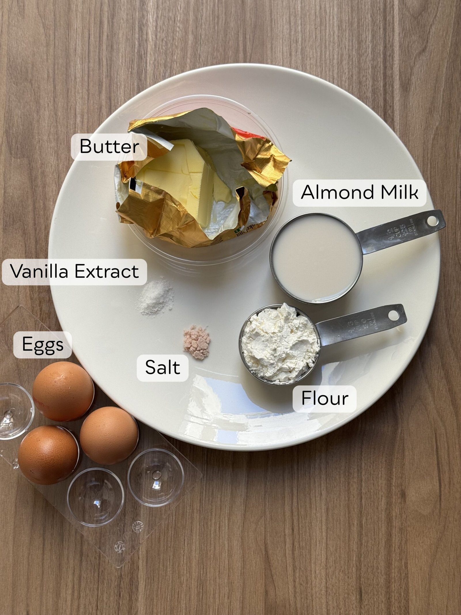 Overhead flat lay of gluten-free Dutch baby pancake ingredients on a white plate, labeled with butter, almond milk, vanilla extract, eggs, salt, and flour
