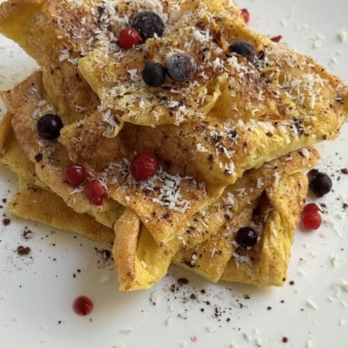 Gluten-free Dutch baby pancake slices stacked on a white plate, topped with desiccated coconut, blueberries, and red currants