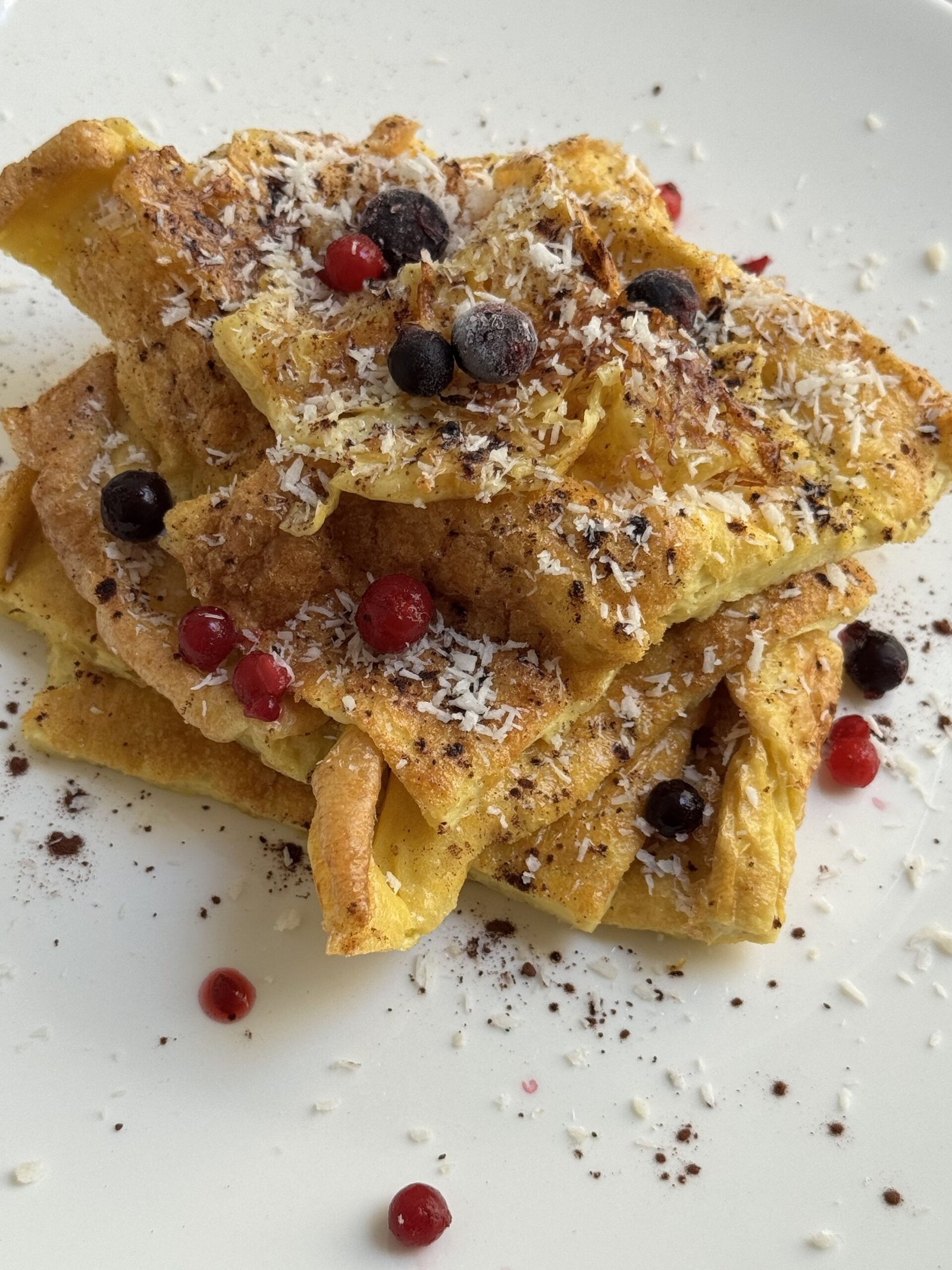 Gluten-free Dutch baby pancake slices stacked on a white plate, topped with desiccated coconut, blueberries, and red currants