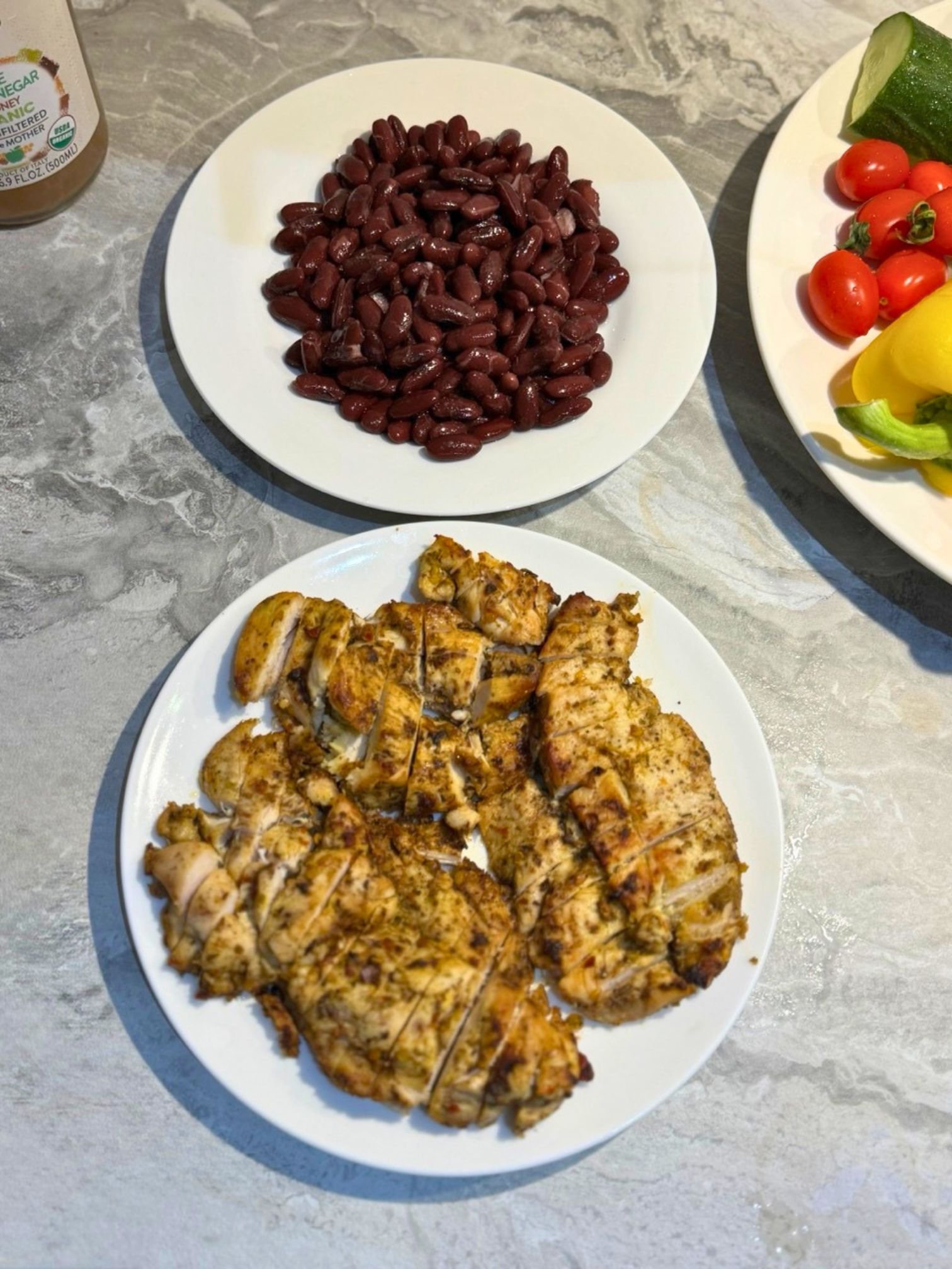 Two white plates resting on a marble countertop; the top plate holds cooked red kidney beans, and the bottom plate holds seasoned, cooked, and sliced chicken breast.
