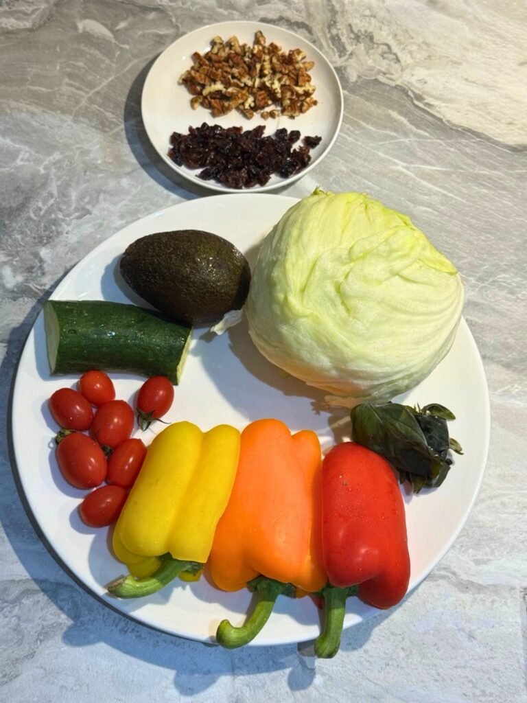 A plate of raw salad ingredients containing an iceberg lettuce head, colorful bell peppers, a cucumber half, cherry tomatoes, an avocado, fresh basil, and a small side plate with chopped nuts and raisins.