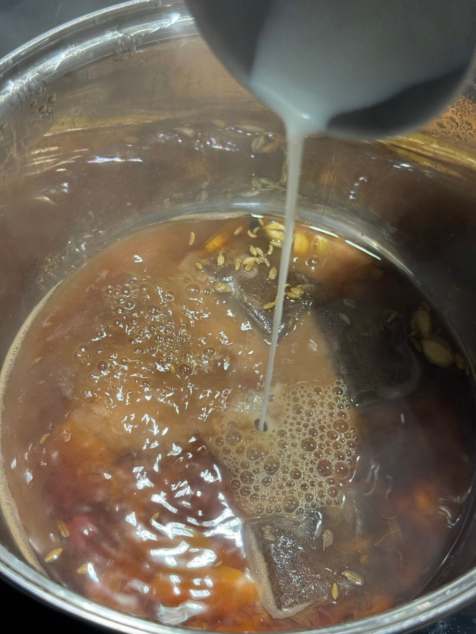 Pouring milk into a saucepan of brewed spiced tea with whole spices and tea bags.