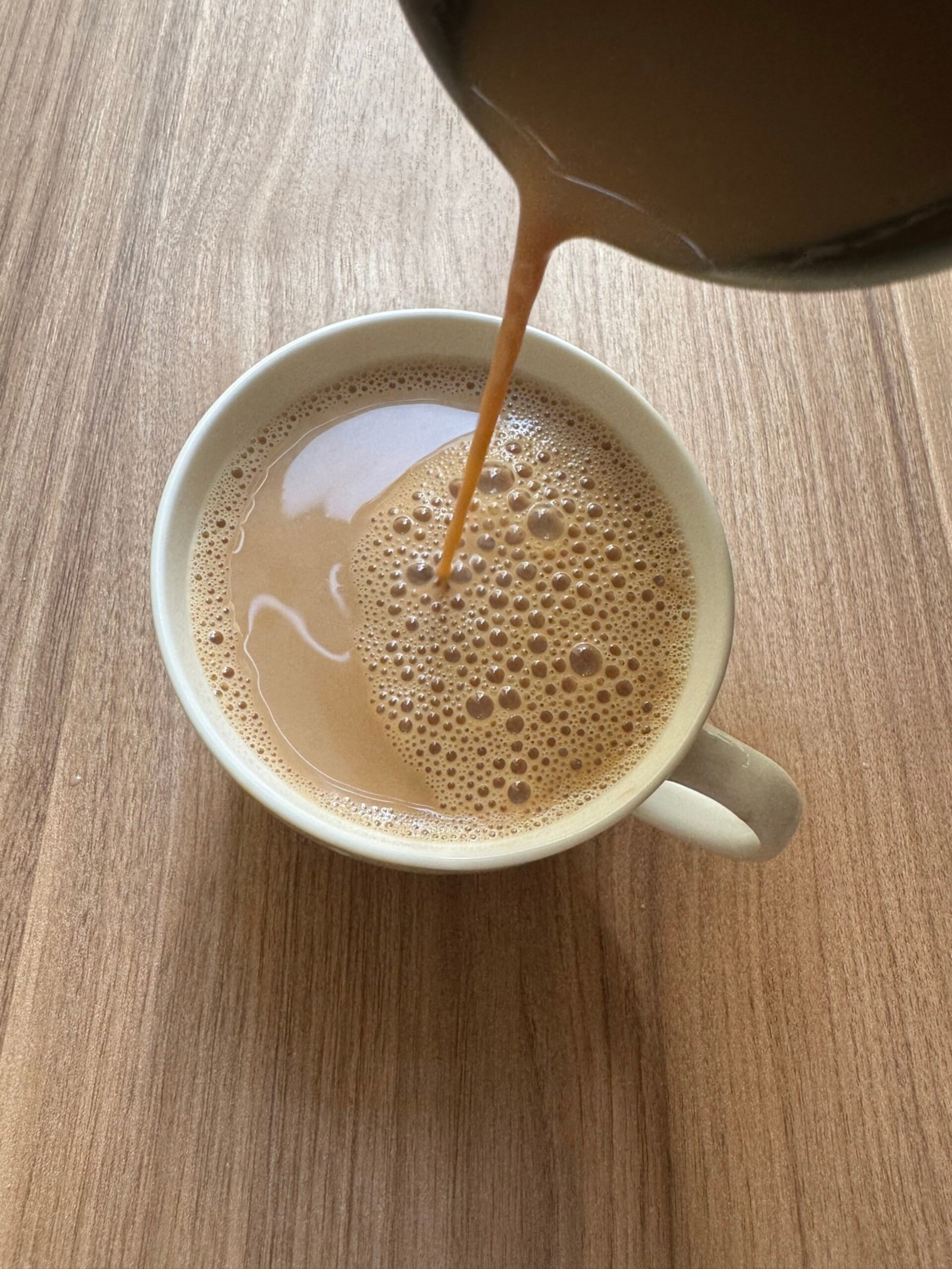 Pouring strained masala chai from a saucepan into a white cup, showing the natural froth on top.