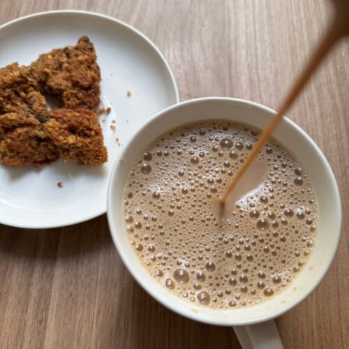 Pouring masala chai into a white cup with frothy bubbles on top, served with a biscuit on the side.