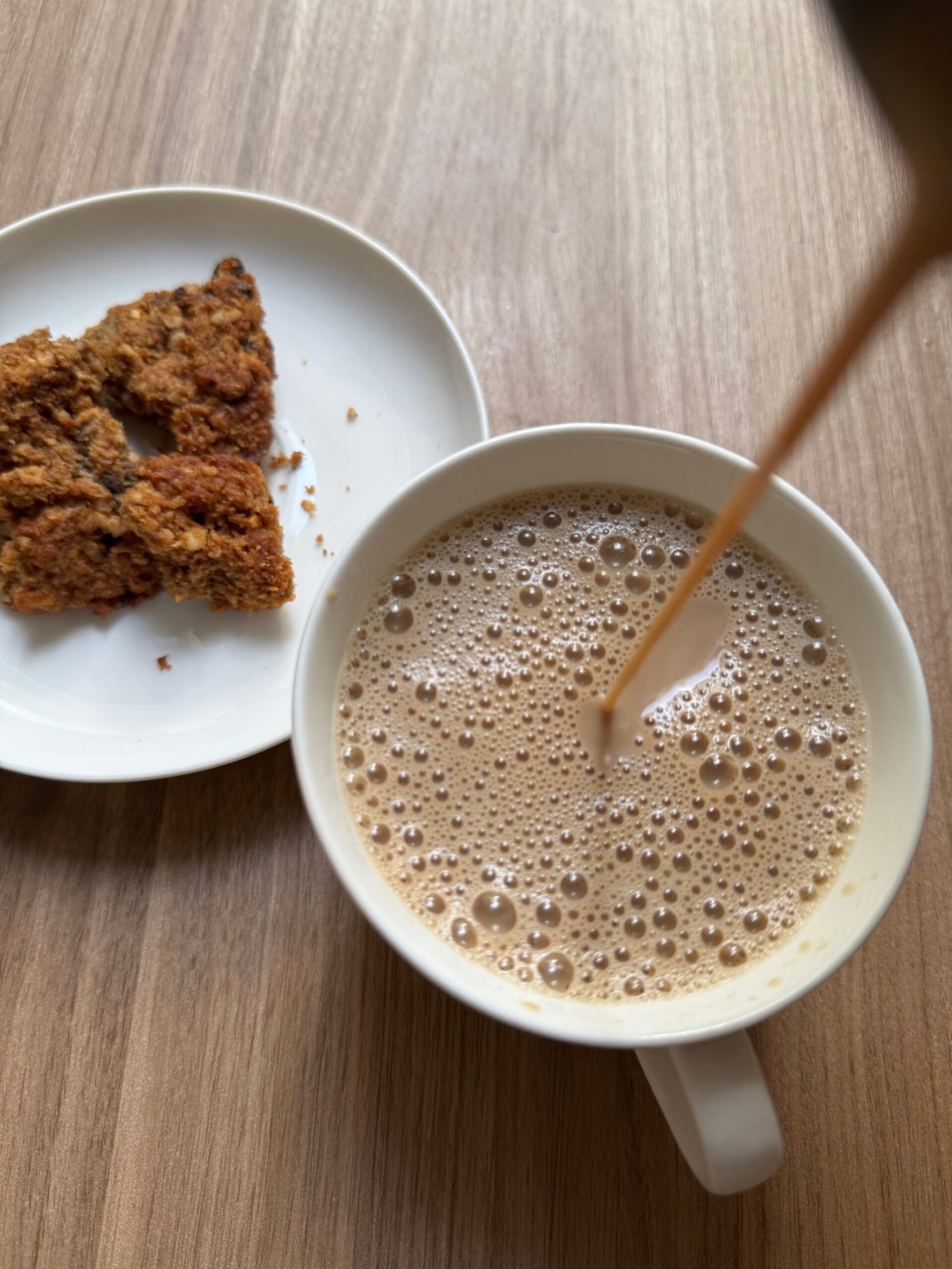 Pouring masala chai into a white cup with frothy bubbles on top, served with a biscuit on the side.