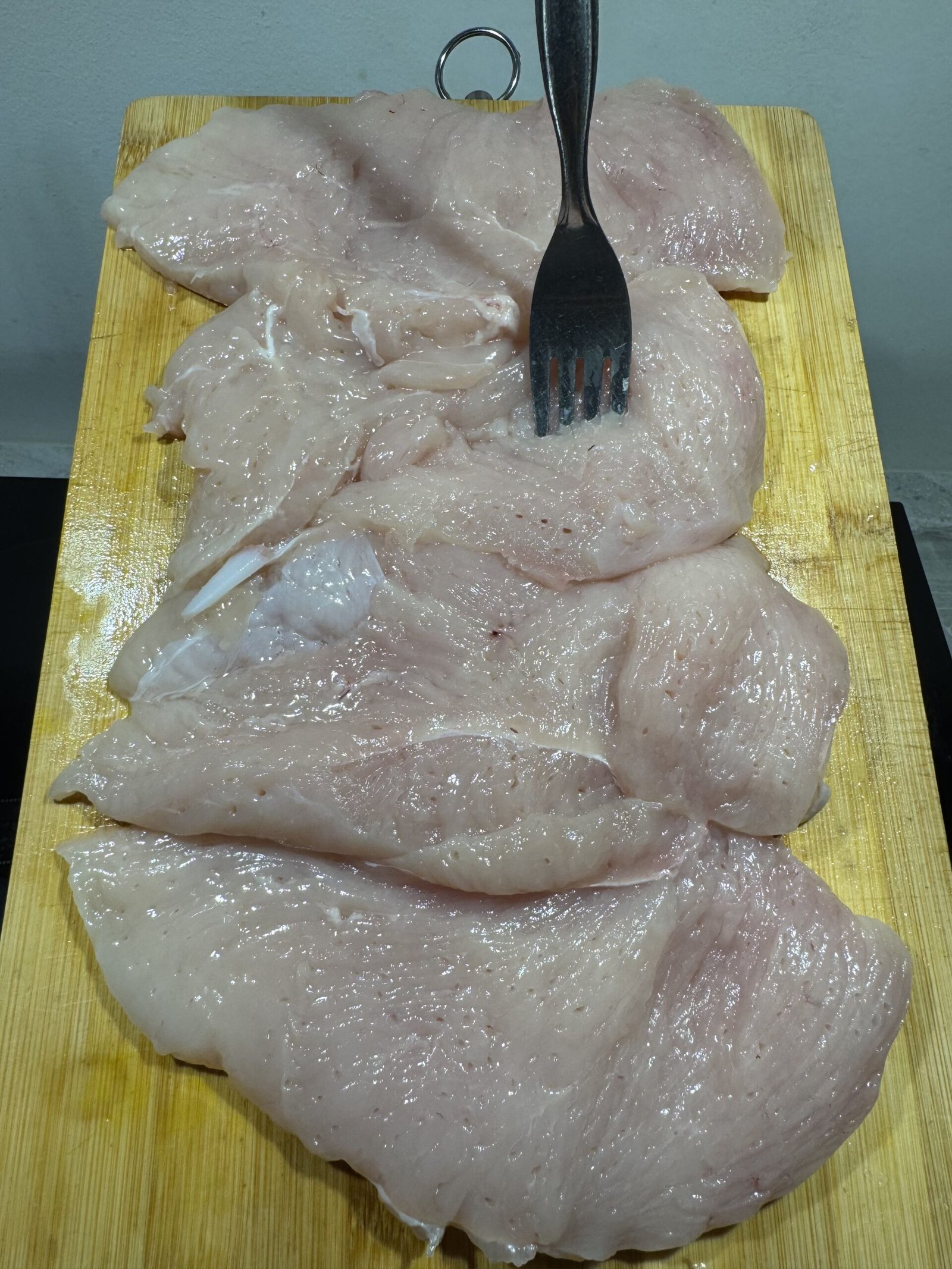 A metal fork piercing one of four raw chicken breasts on a wooden cutting board to tenderize the meat.
