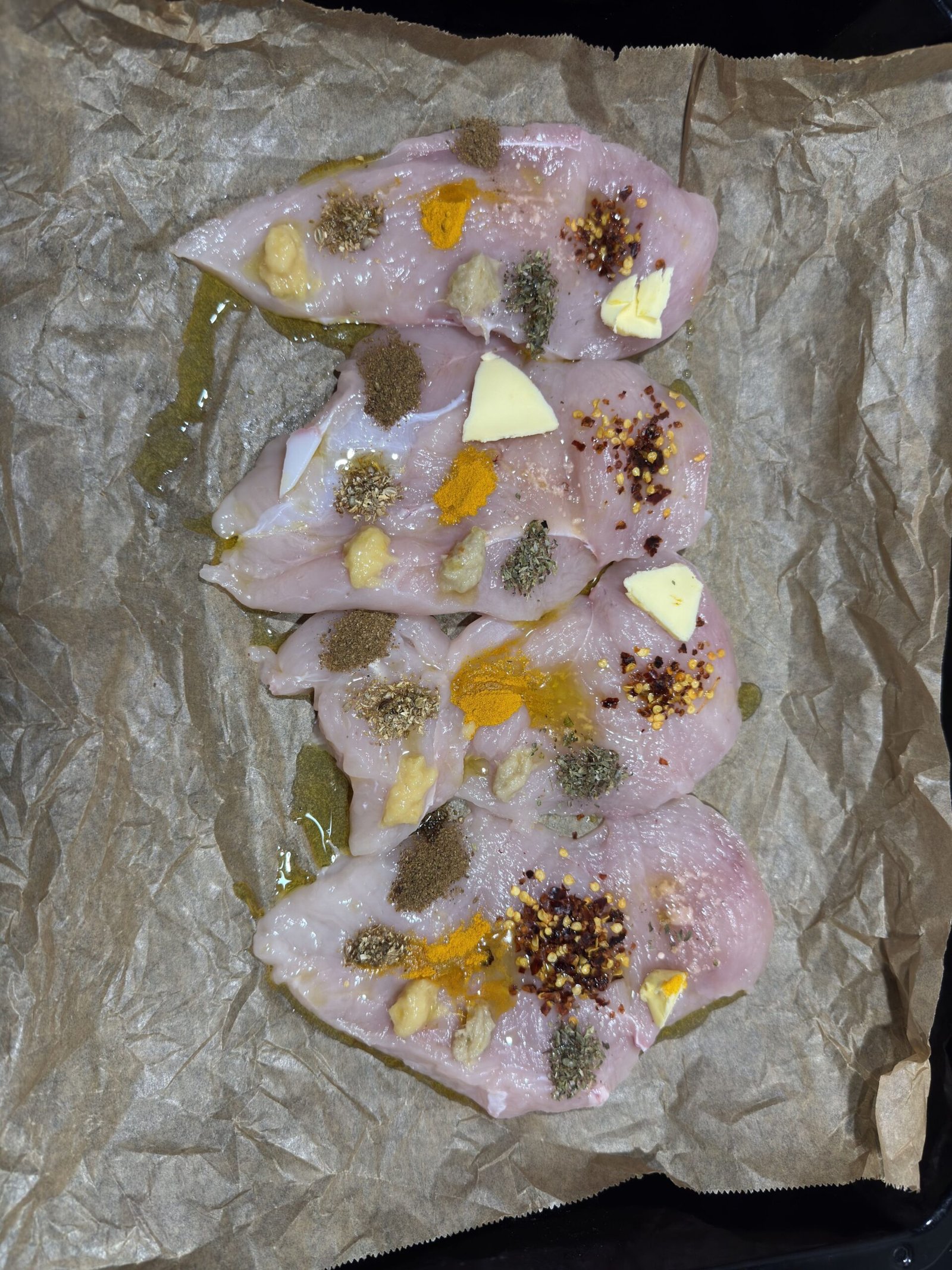 Four raw chicken breasts lined up on brown parchment paper, dotted with small pats of butter, oil, minced garlic, and distinct piles of assorted colorful dry spices before being rubbed in.