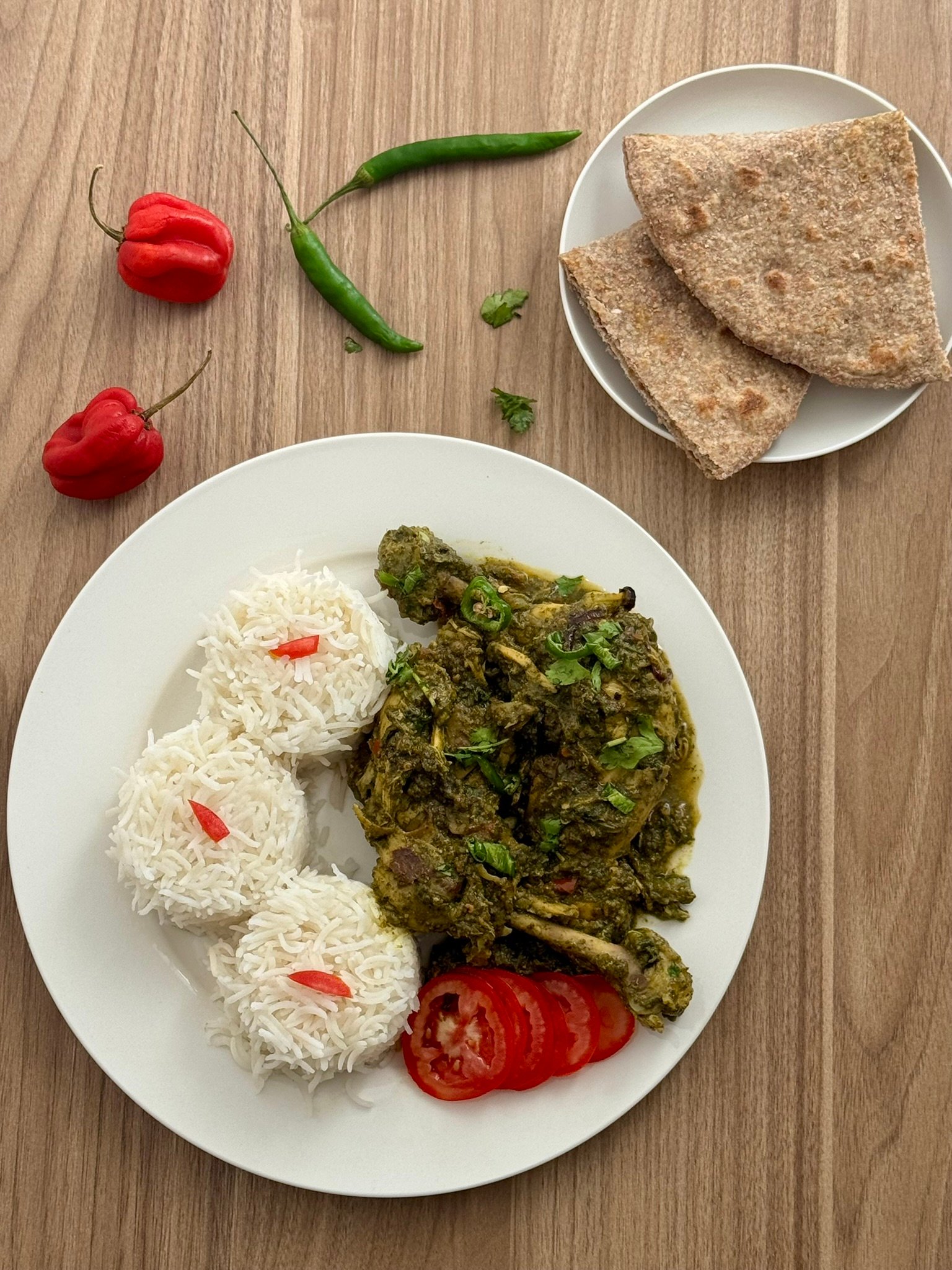 Overhead view of palak chicken plated with steamed basmati rice, sliced tomatoes, fresh green chilies, and whole wheat roti on the side.