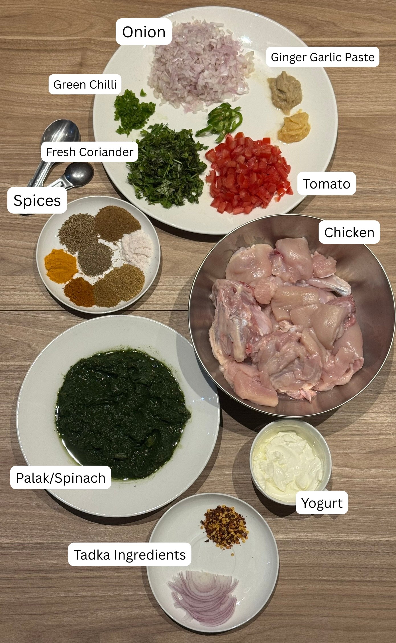 All ingredients for palak chicken laid out and labeled, including chopped onion, ginger garlic paste, green chilies, fresh coriander, tomato, spices, raw chicken pieces, blanched spinach, yogurt, and tadka ingredients.
