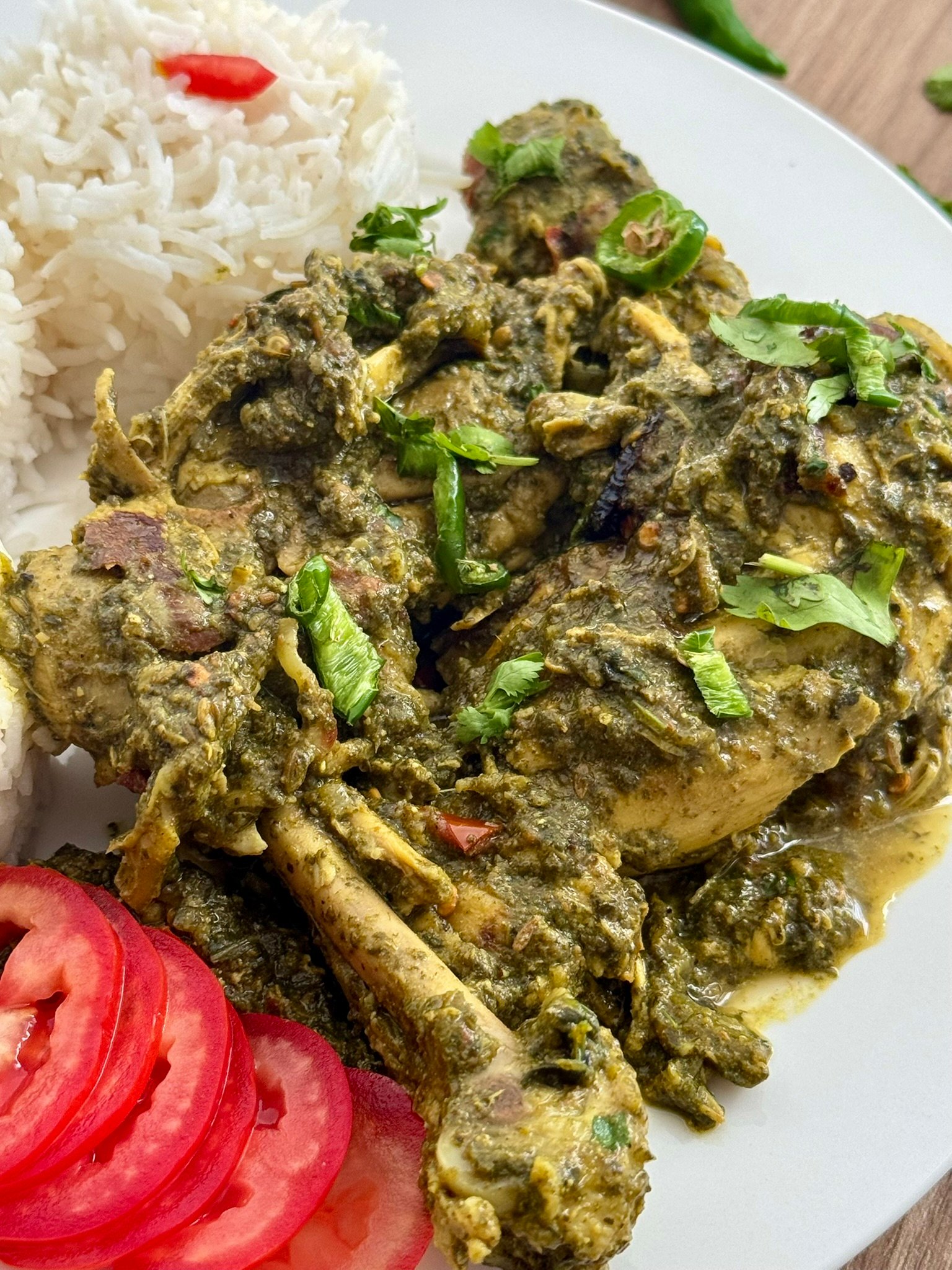 Close-up of bone-in palak chicken pieces coated in thick spinach gravy, garnished with fresh cilantro and sliced green chilies.