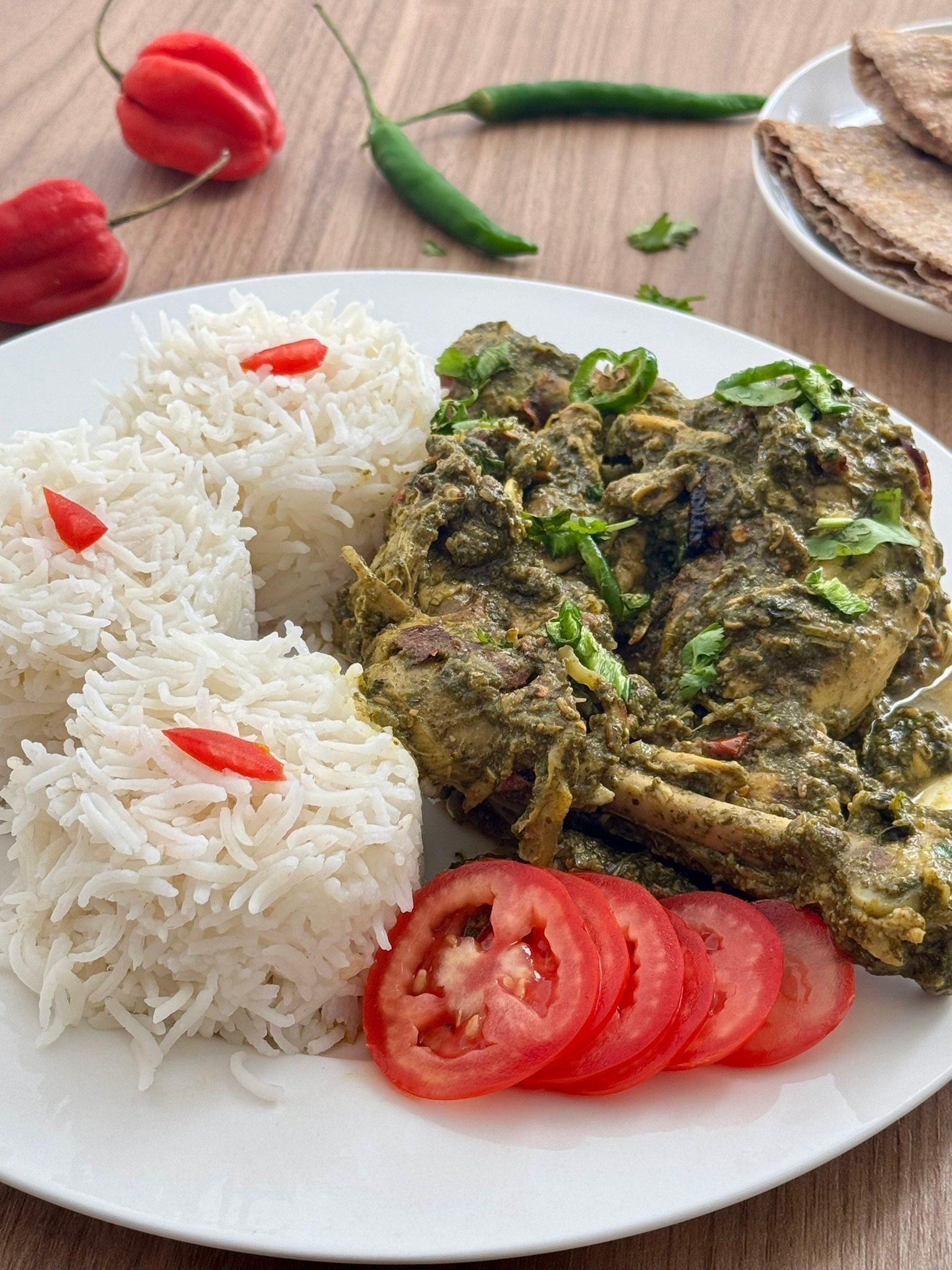 Palak chicken served on a white plate with steamed basmati rice, sliced tomatoes, fresh green chilies, and roti on the side.