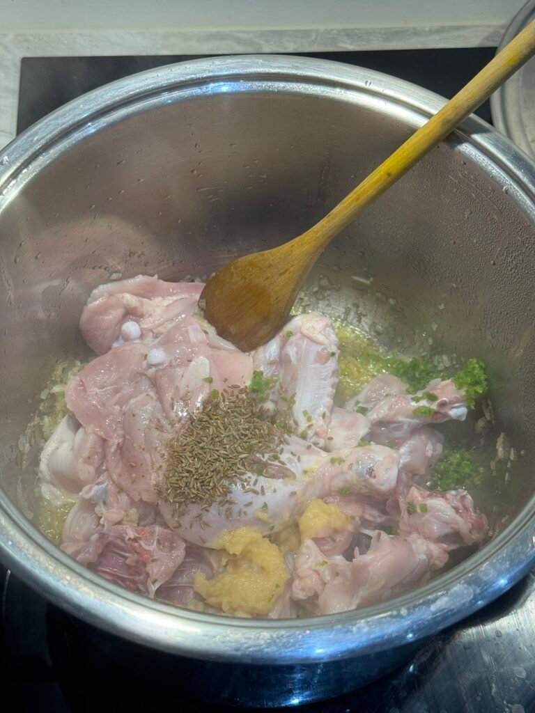 Raw bone-in chicken pieces added to the pan with cumin seeds, ginger garlic paste, and a wooden spoon.