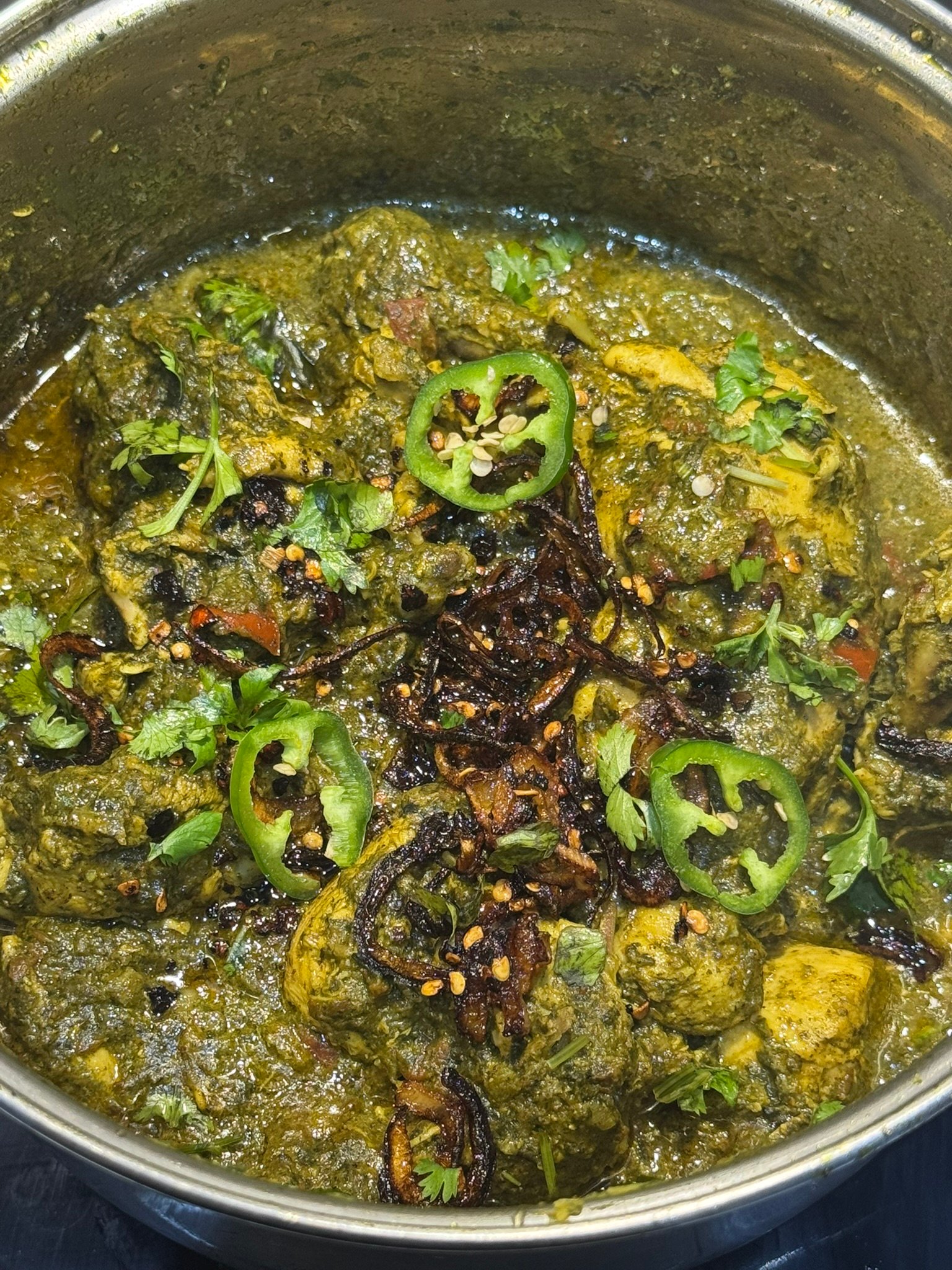Finished palak chicken in the pot with the ghee tarka poured on top, showing crispy fried onions, red chili flakes, sliced green chilies, and fresh cilantro.