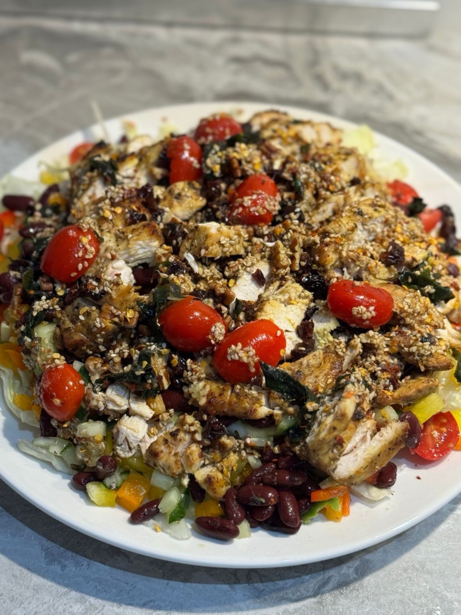 A detailed close-up shot of a tossed chicken and kidney bean salad, highlighting the textures of the sliced chicken, glossy cherry tomatoes, mixed greens, and sprinkled sesame seeds.