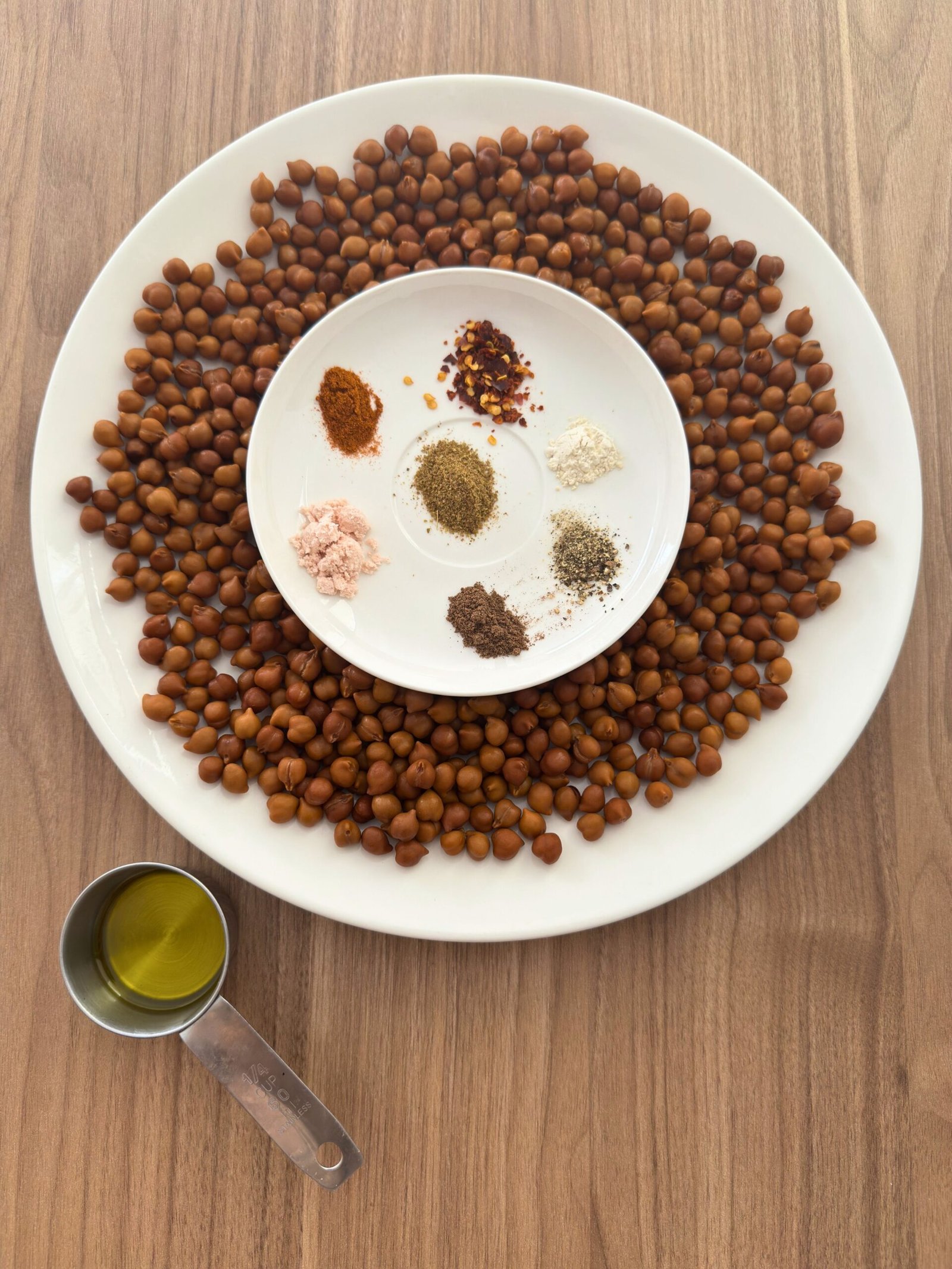 Overhead flat-lay of ingredients for roasted chickpeas: boiled brown chickpeas arranged in a circle on a white plate, with a center bowl containing seven measured spice piles (chilli flakes, cayenne pepper, black pepper, pink Himalayan salt, garam masala, ground allspice, and garlic powder), and a measuring cup of olive oil to the side