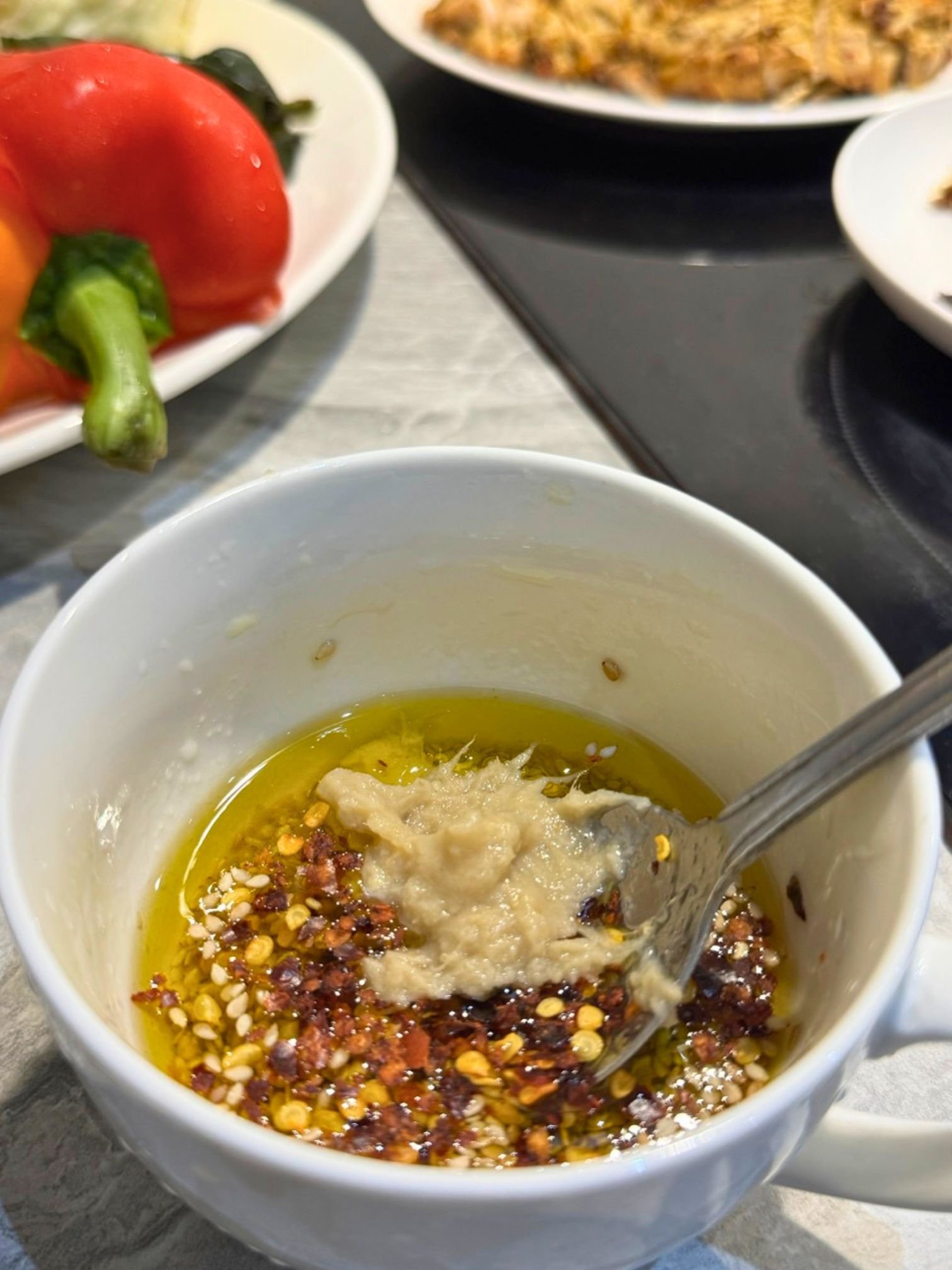 A close-up view inside a white mug showing the unmixed ingredients for a salad dressing, featuring olive oil, a scoop of garlic paste, red chili flakes, and sesame seeds with a spoon resting inside.