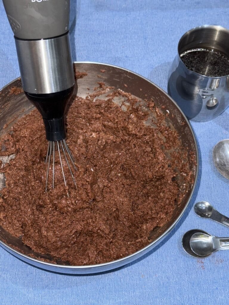 Thick gluten-free chocolate cake batter being mixed with an immersion blender whisk attachment, with a cup of brewed coffee beside the bowl.