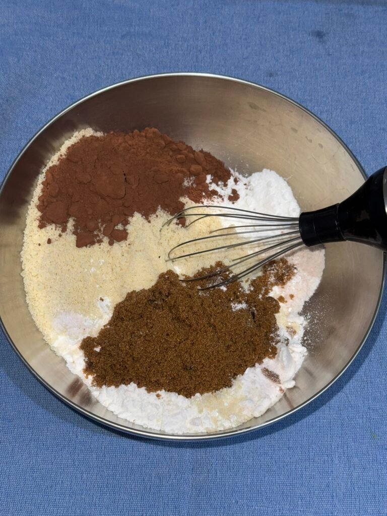 Cocoa powder, almond flour, brown sugar, and other dry ingredients mounded separately in a metal mixing bowl before being whisked together