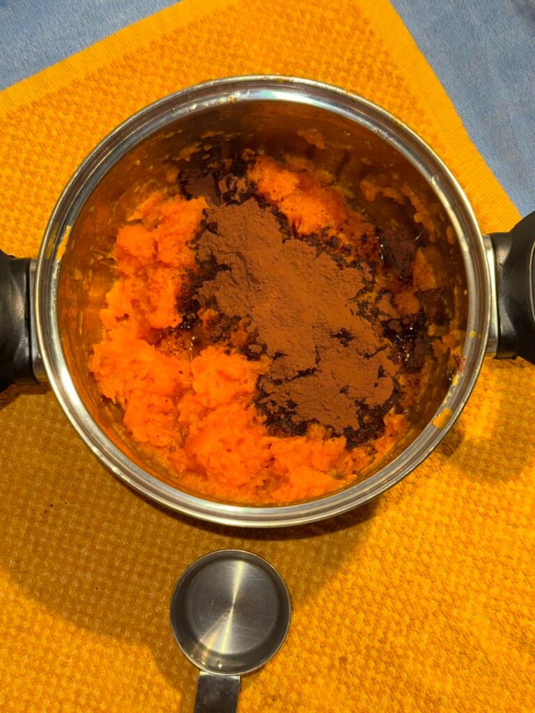 Mashed sweet potato in a metal mixing bowl topped with cocoa powder and vanilla extract, ready to be blended into dairy-free chocolate frosting.