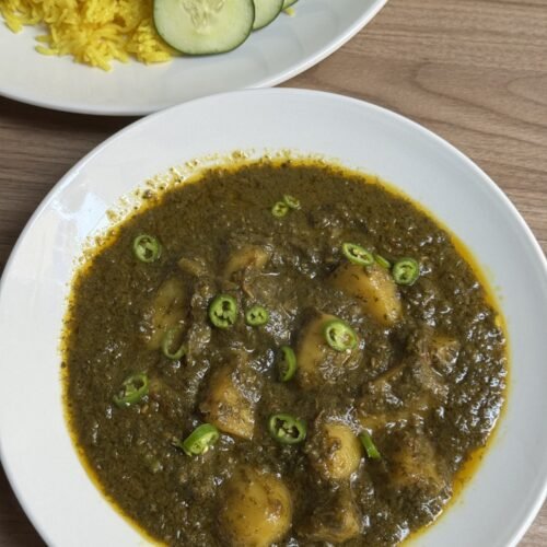 Aloo palak served in a white bowl with yellow rice and cucumber slices on the side