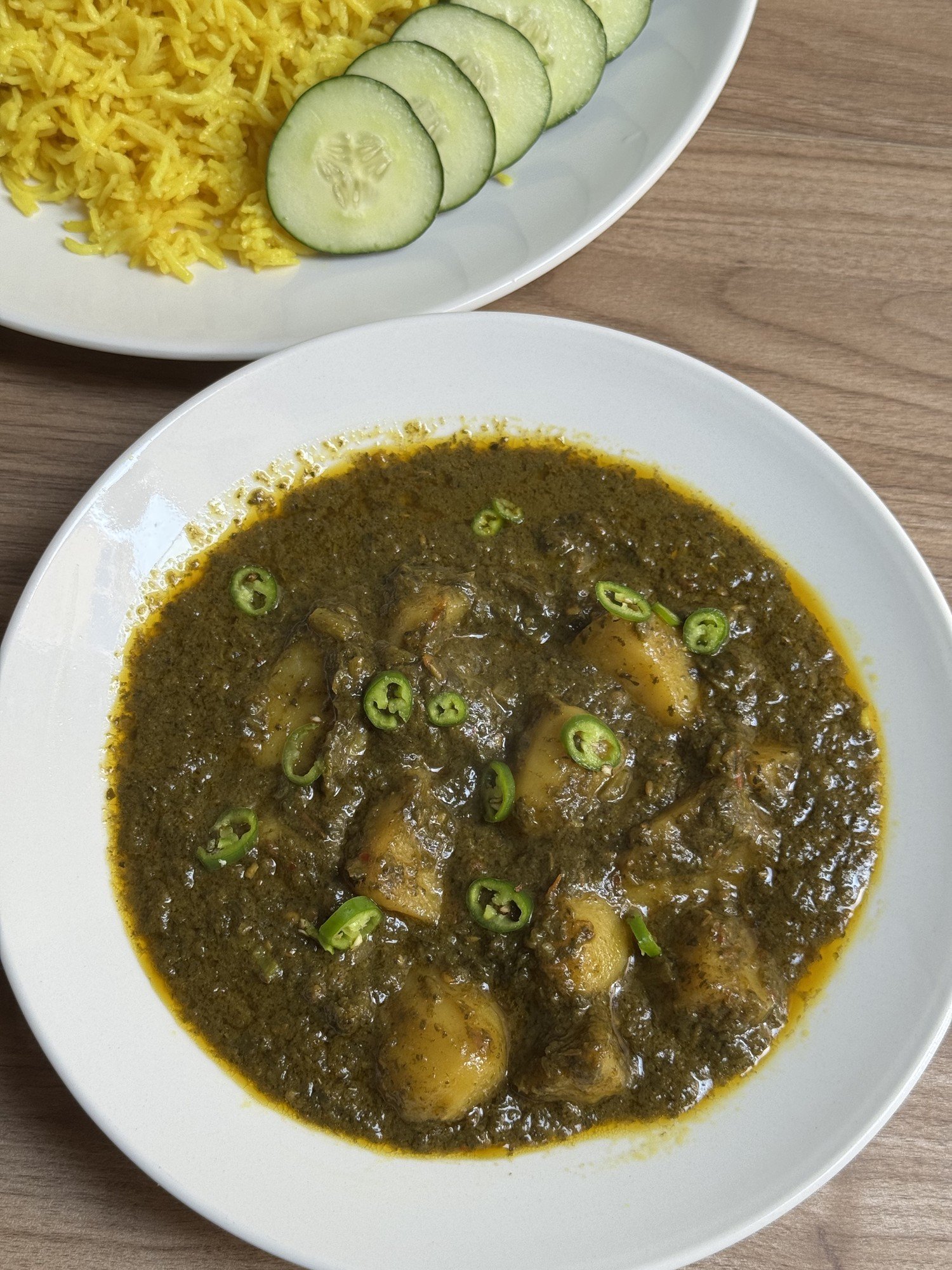Aloo palak served in a white bowl with yellow rice and cucumber slices on the side